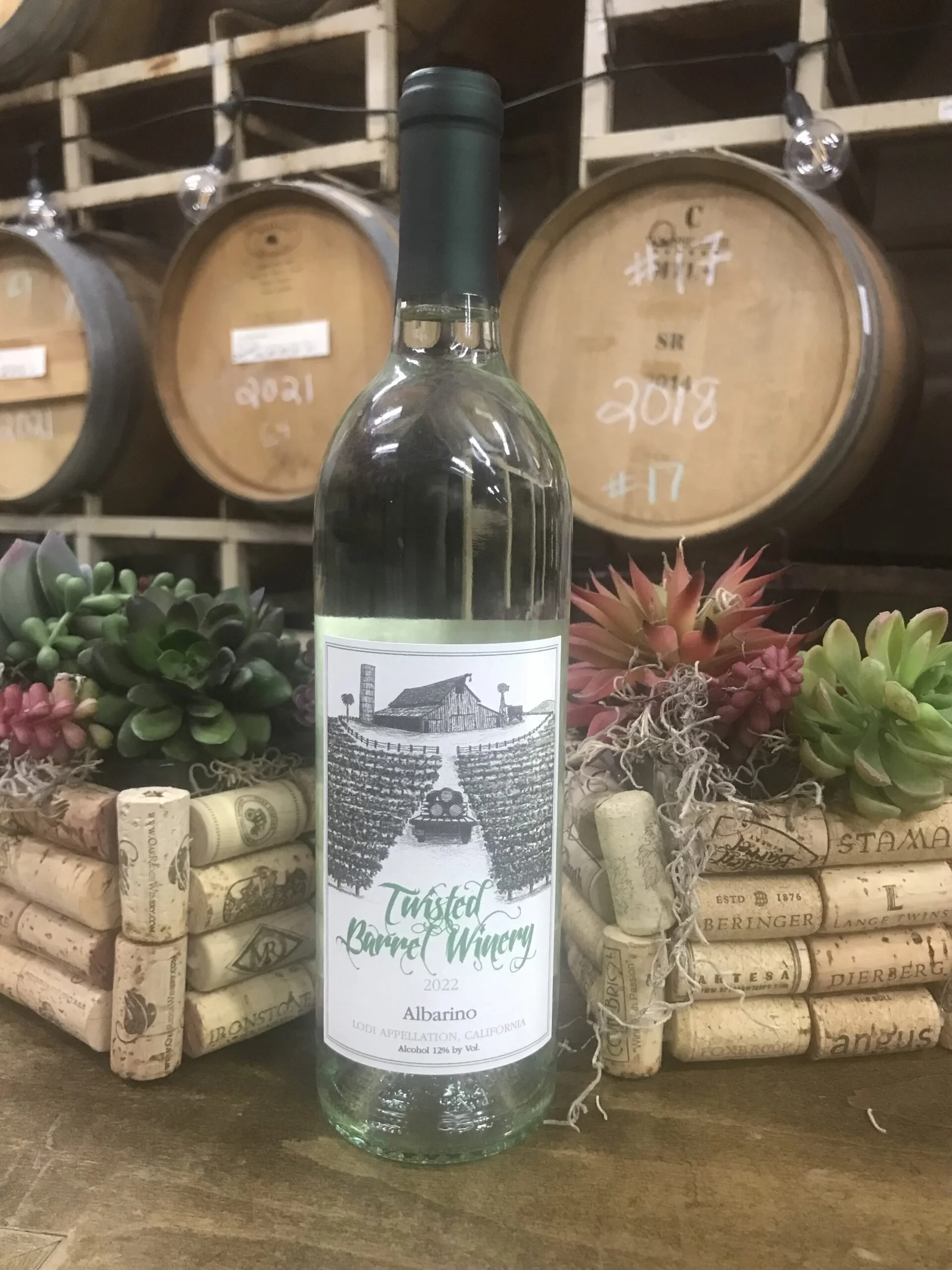 A bottle of Twisted Barrel Winery Albarino wine placed on a wooden surface, with wine barrels in the background and decorative succulents and corks around it.