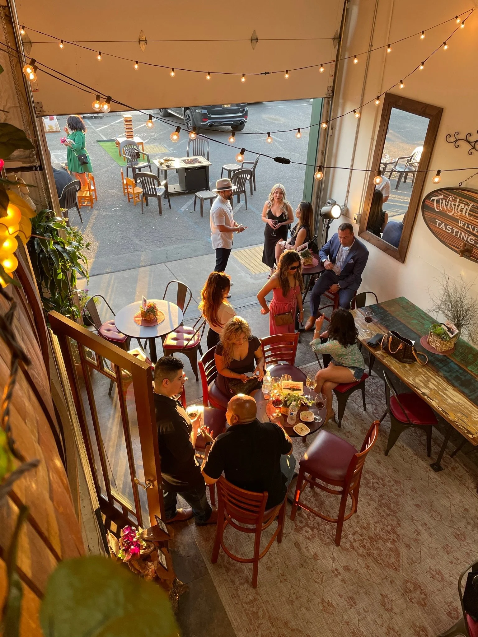 People dining inside a cozy restaurant with string lights, with others standing outside near parked cars.