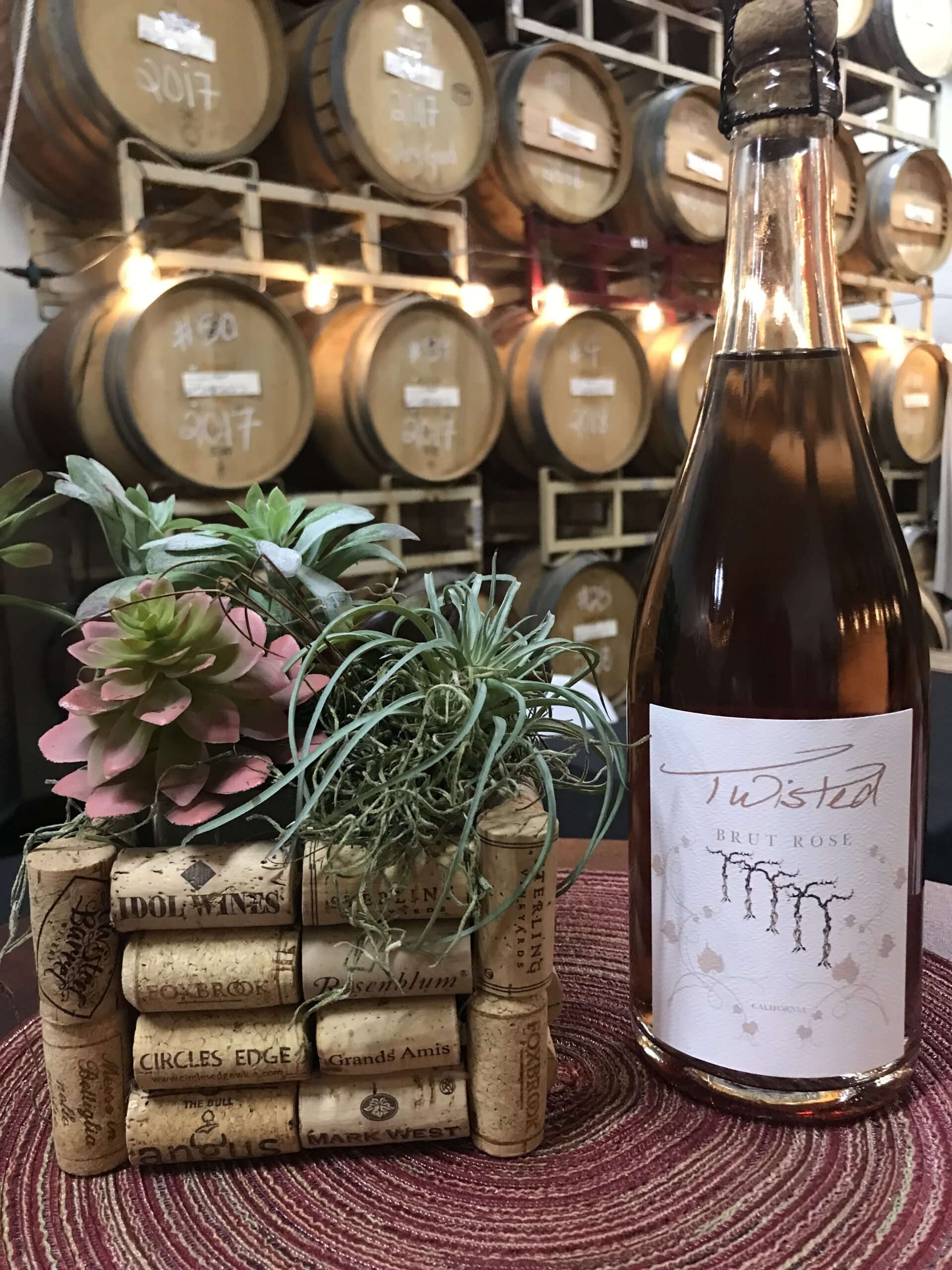 A wine bottle labeled 'Twisted' Brut Rose with a plant and corks in front, set on a red woven placemat, with wine barrels in the background.