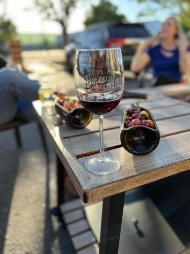 A glass of red wine on a wooden table outdoors, with wine bottles lying on their sides and a woman in the background smiling and talking.