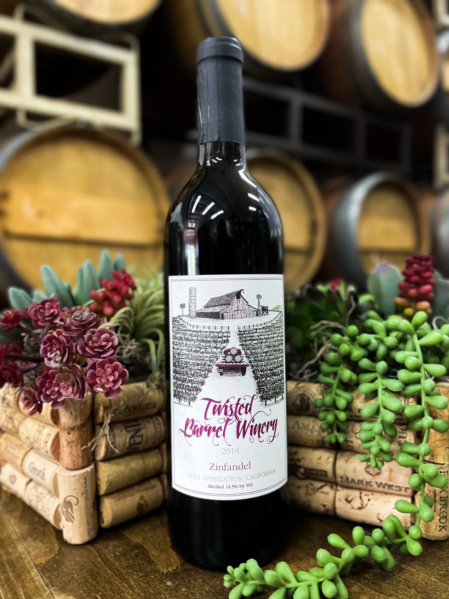 A bottle of Twisted Barrel Winery Zinfandel wine from 2018 placed on a wooden surface surrounded by succulents and wine corks, with wooden wine barrels in the background.