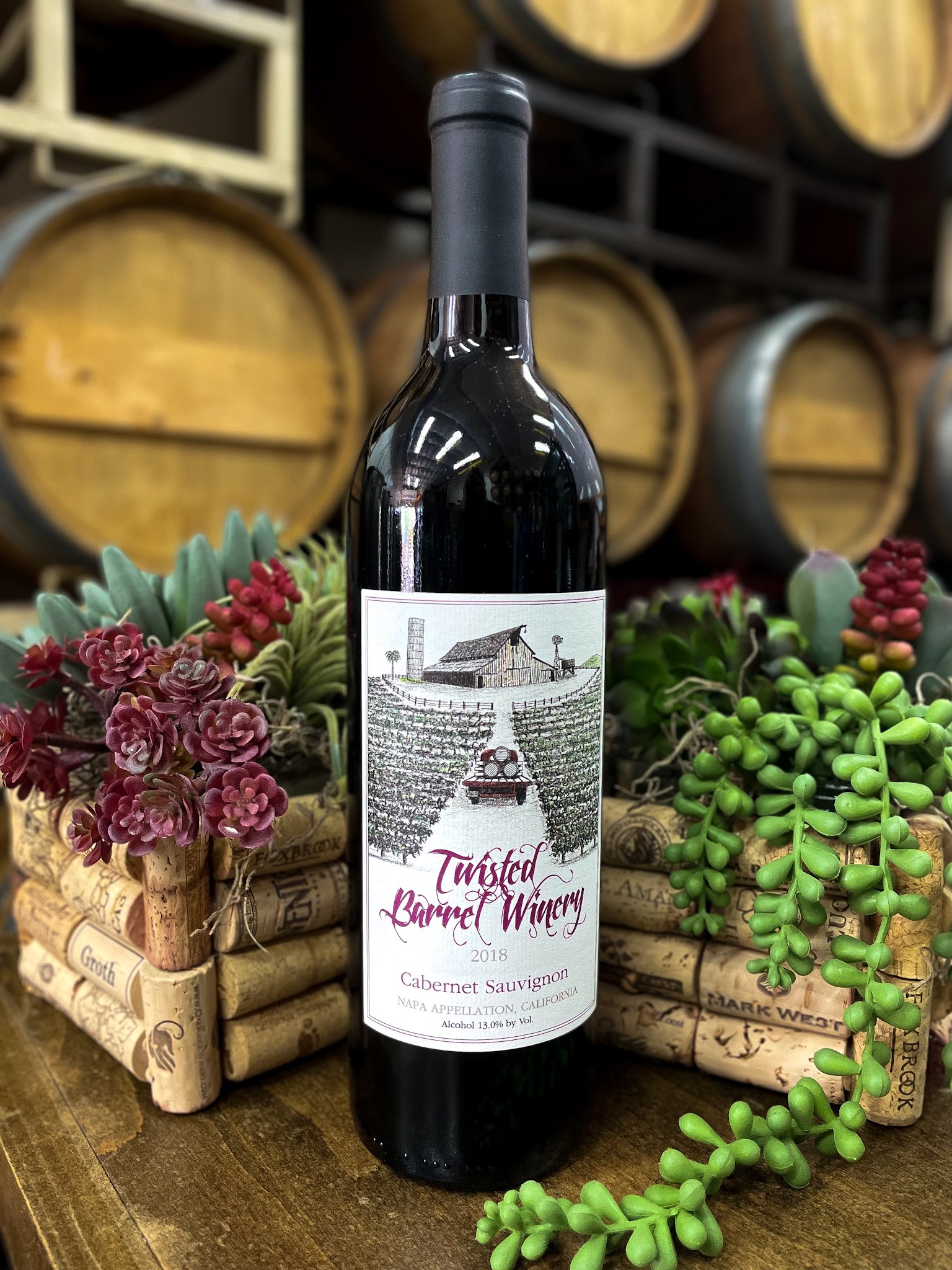 A bottle of Twisted Barre Winery 2018 Cabernet Sauvignon placed on a wooden surface, surrounded by small plant arrangements with purple and green succulent-like plants, with wine barrels in the background.