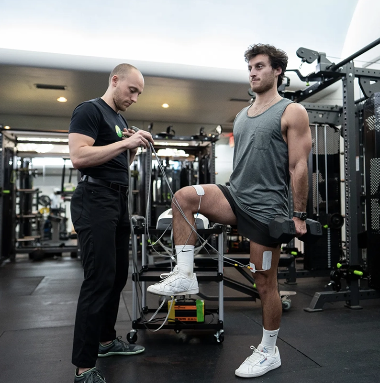 A man performs a single-leg squat with weights in a gym, monitored by a trainer.