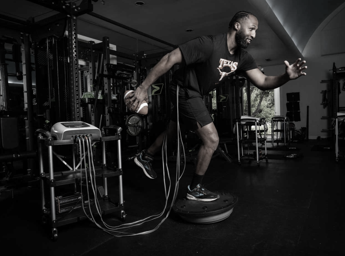 Man exercising in a gym on a balance platform, holding a football, with gym equipment in the background.