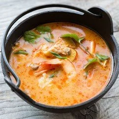 A black bowl of spicy seafood soup garnished with fresh herbs on a gray wooden surface.