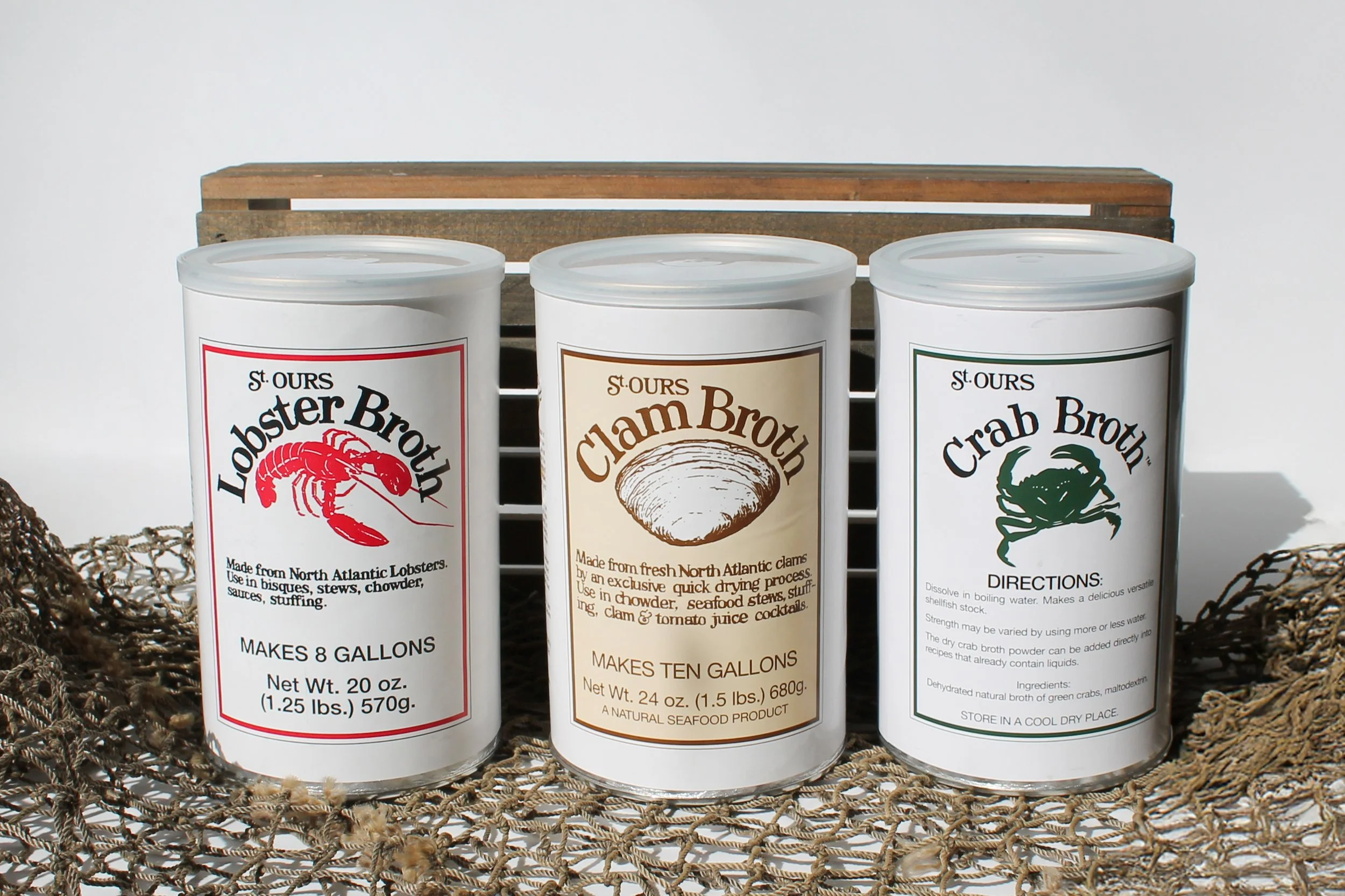 Three cylindrical containers of seafood broth on a woven mat with a wooden crate in the background. The left container is labeled 'Lobster Broth,' the middle 'Clam Broth,' and the right 'Crab Broth.'