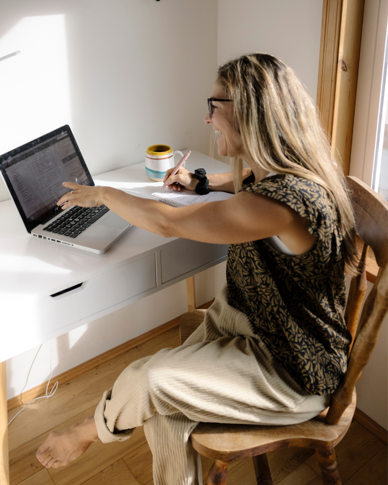 A woman sitting at a white desk, smiling and using a laptop while writing in a notebook, with a mug beside her, near a window with sunlight streaming in.