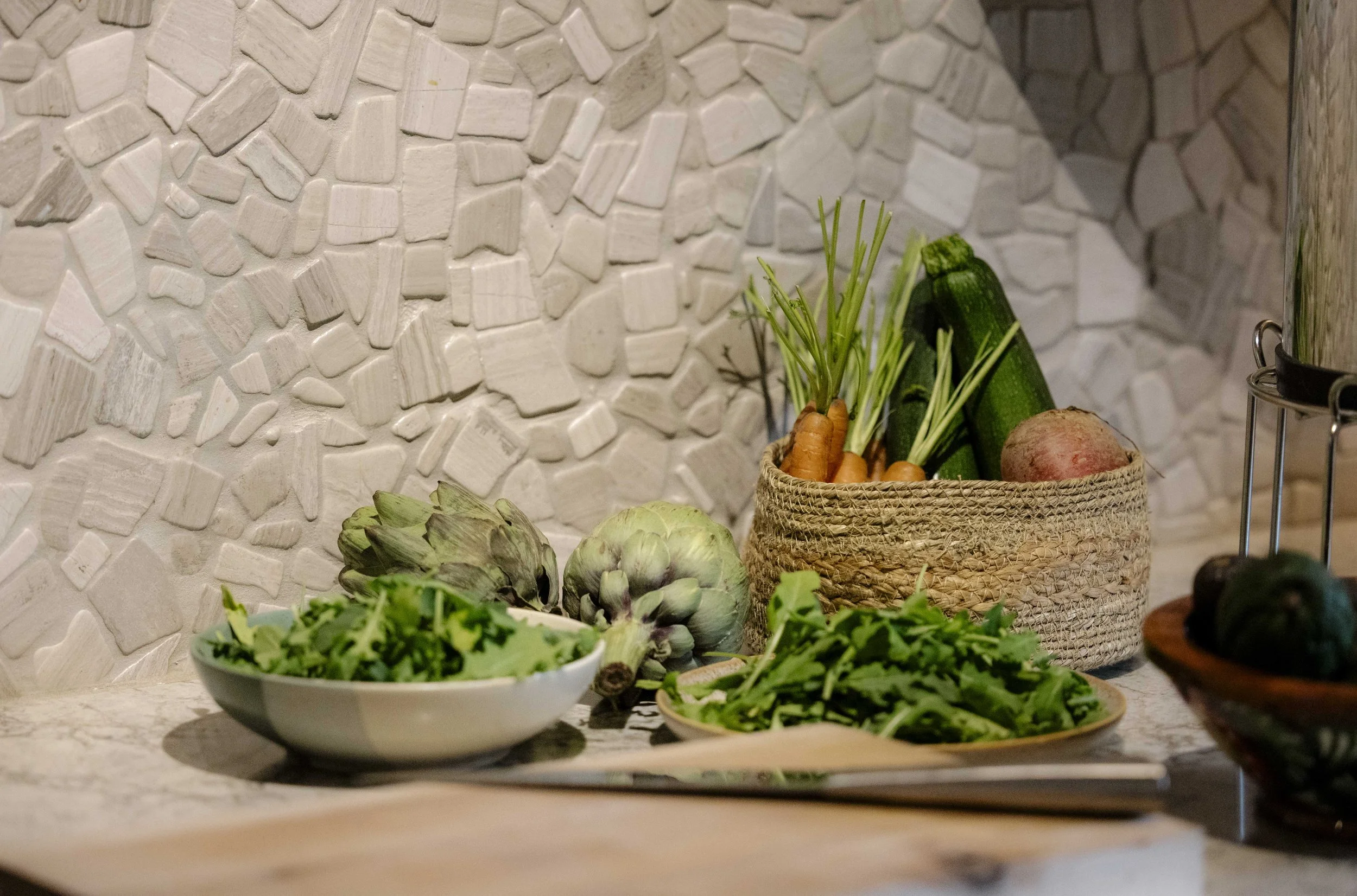 Fresh vegetables and greens on a kitchen counter, including carrots with greens, artichokes, zucchini, and lettuce, with a textured white backsplash in the background.