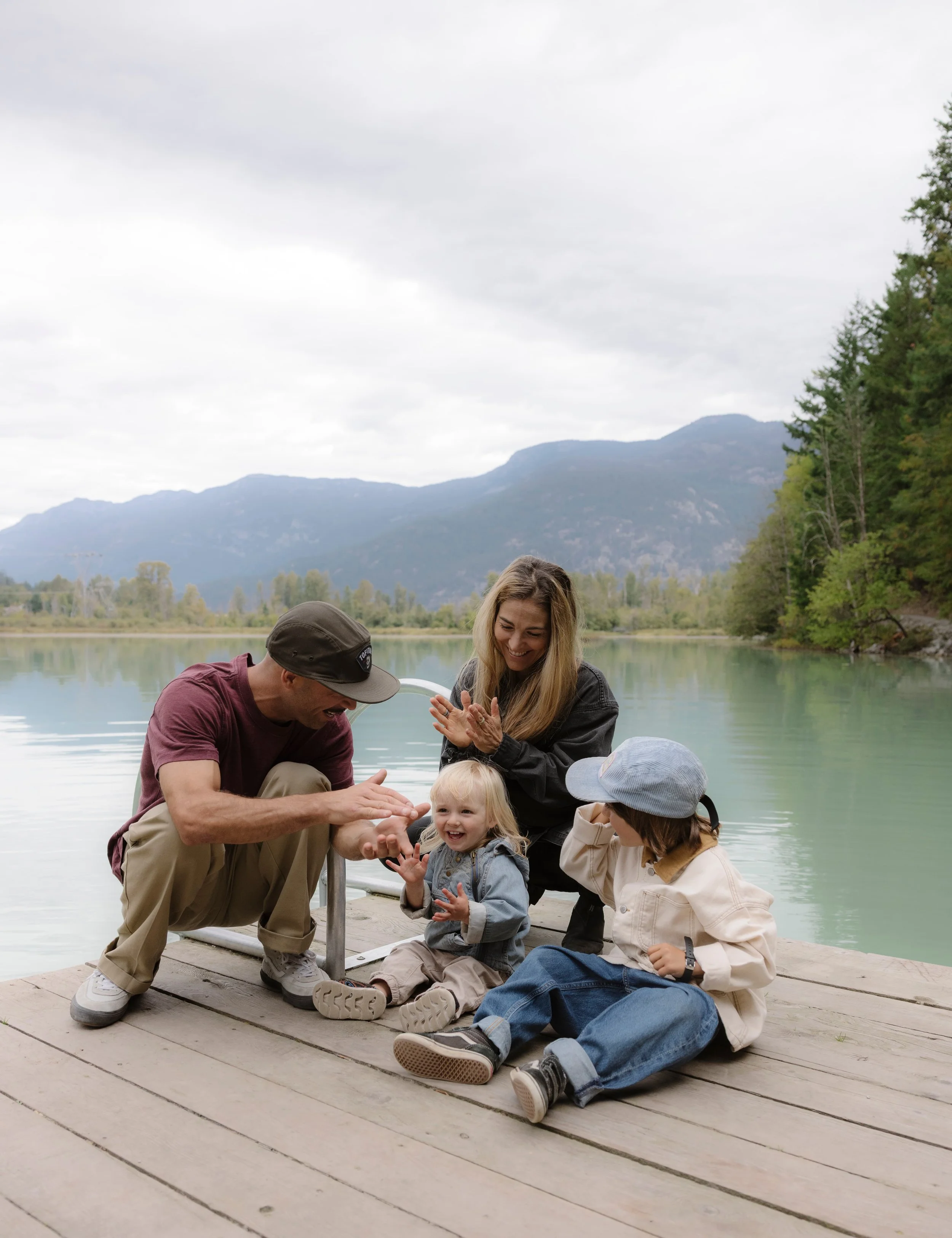 A family enjoying time together on a wooden dock by a lake, surrounded by mountains and trees under a cloudy sky.