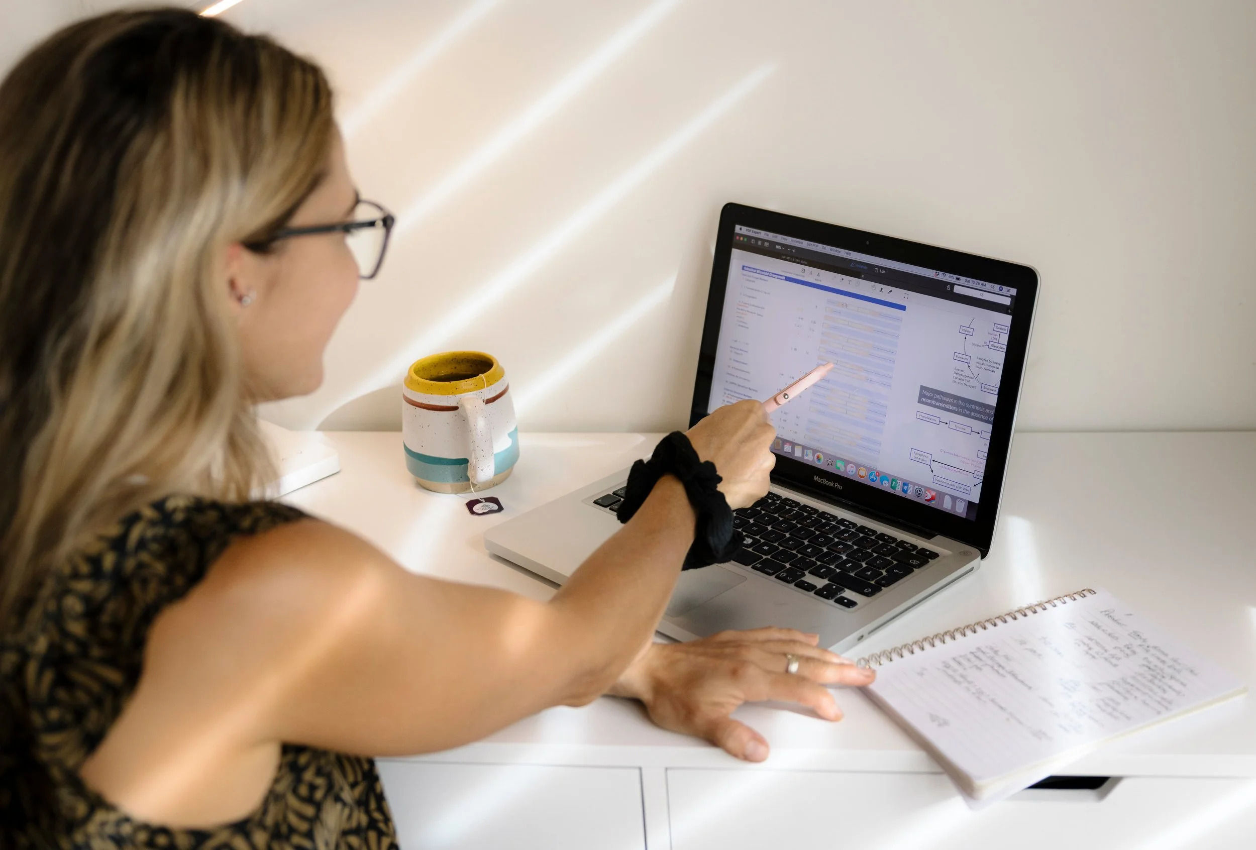 Woman with glasses working on a laptop at a white desk, pointing at the screen, with a coffee mug and notebook nearby.