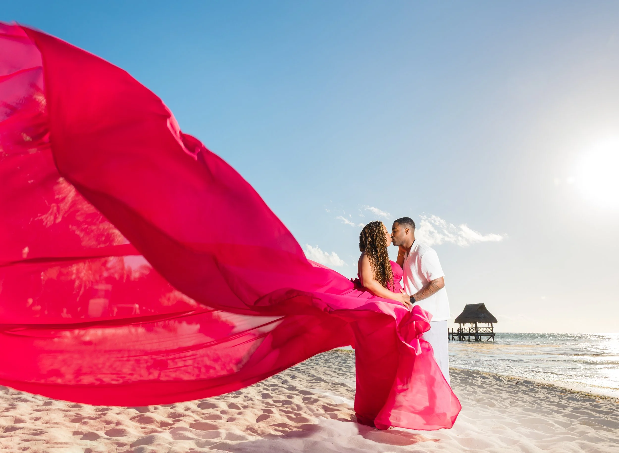 A couple standing close together on a beach during sunset, with a woman wearing a flowing red dress and a man in black, near a wooden pier with a thatched hut in the background.