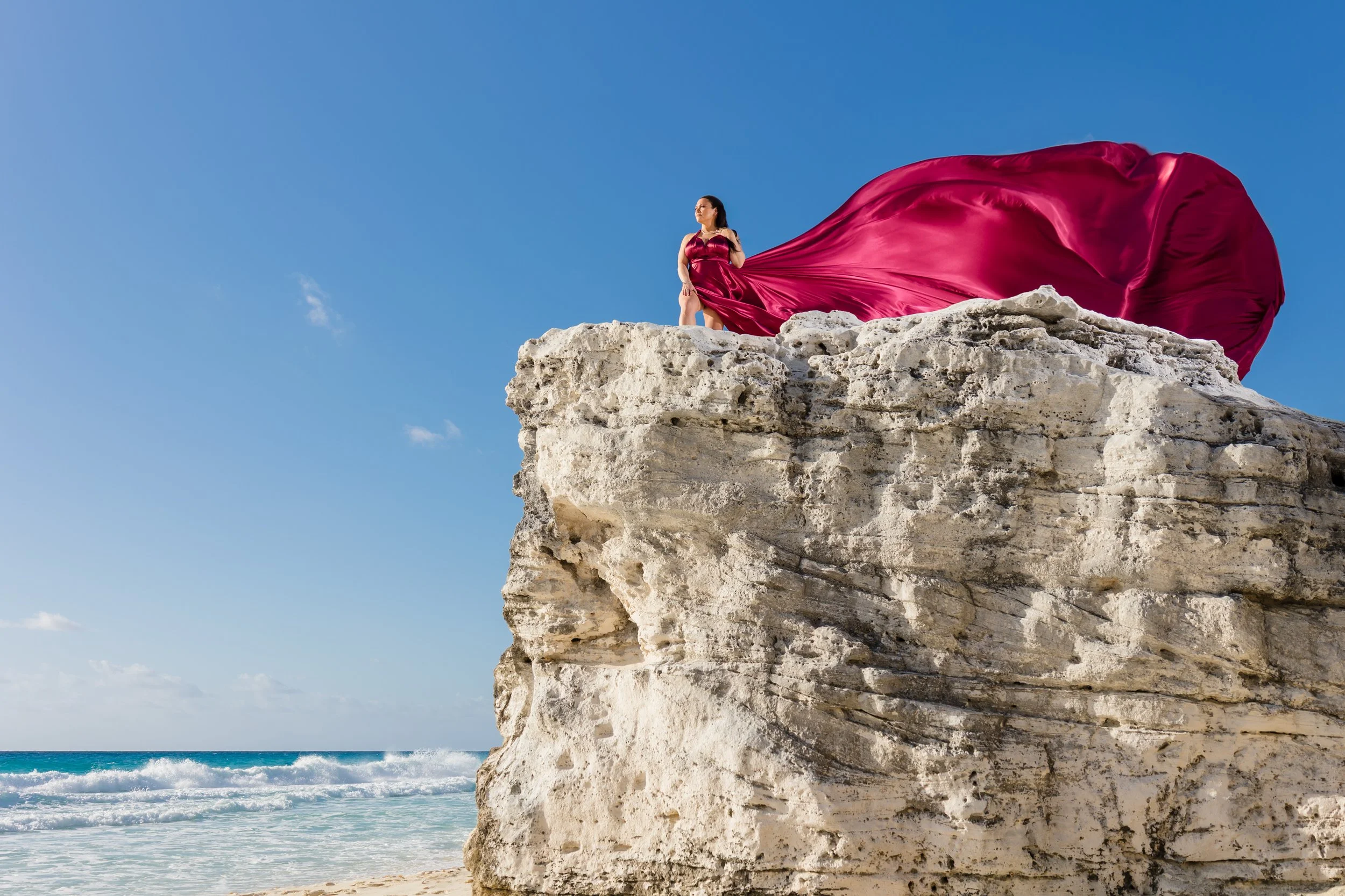 A couple standing on a rocky beach, the woman in a red gown with a long train and the man shirtless with red shorts, overlooking the ocean.
