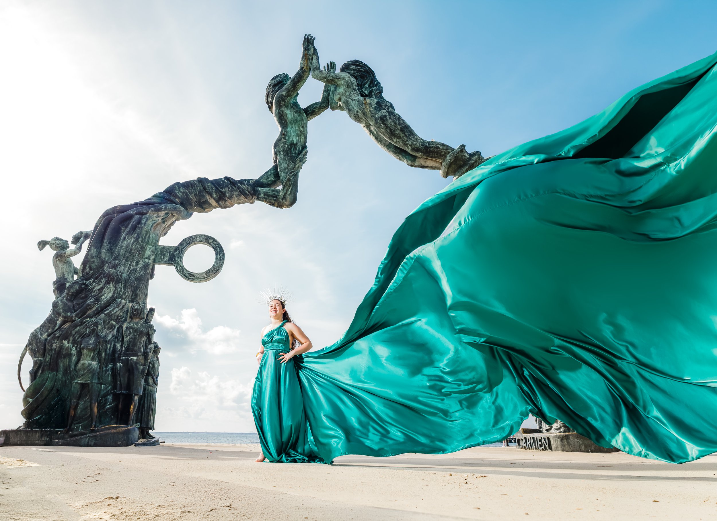 A woman in a black dress poses in front of a large sculpture of two intertwined figures, with palm trees and a pier in the background, on a cloudy day at the beach.