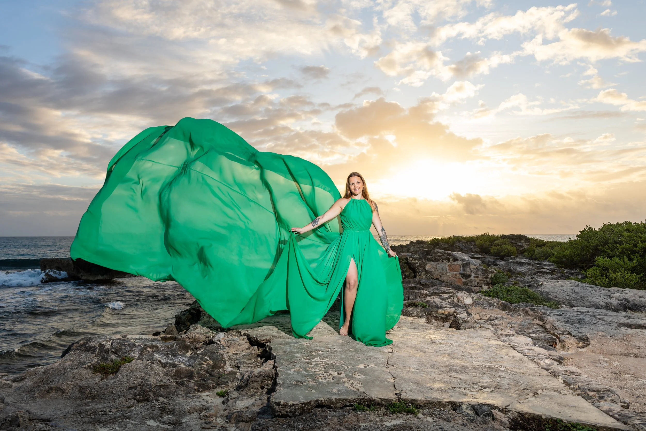 A couple stands on a rocky beach at sunset, with a large green fabric flowing in the wind behind them.