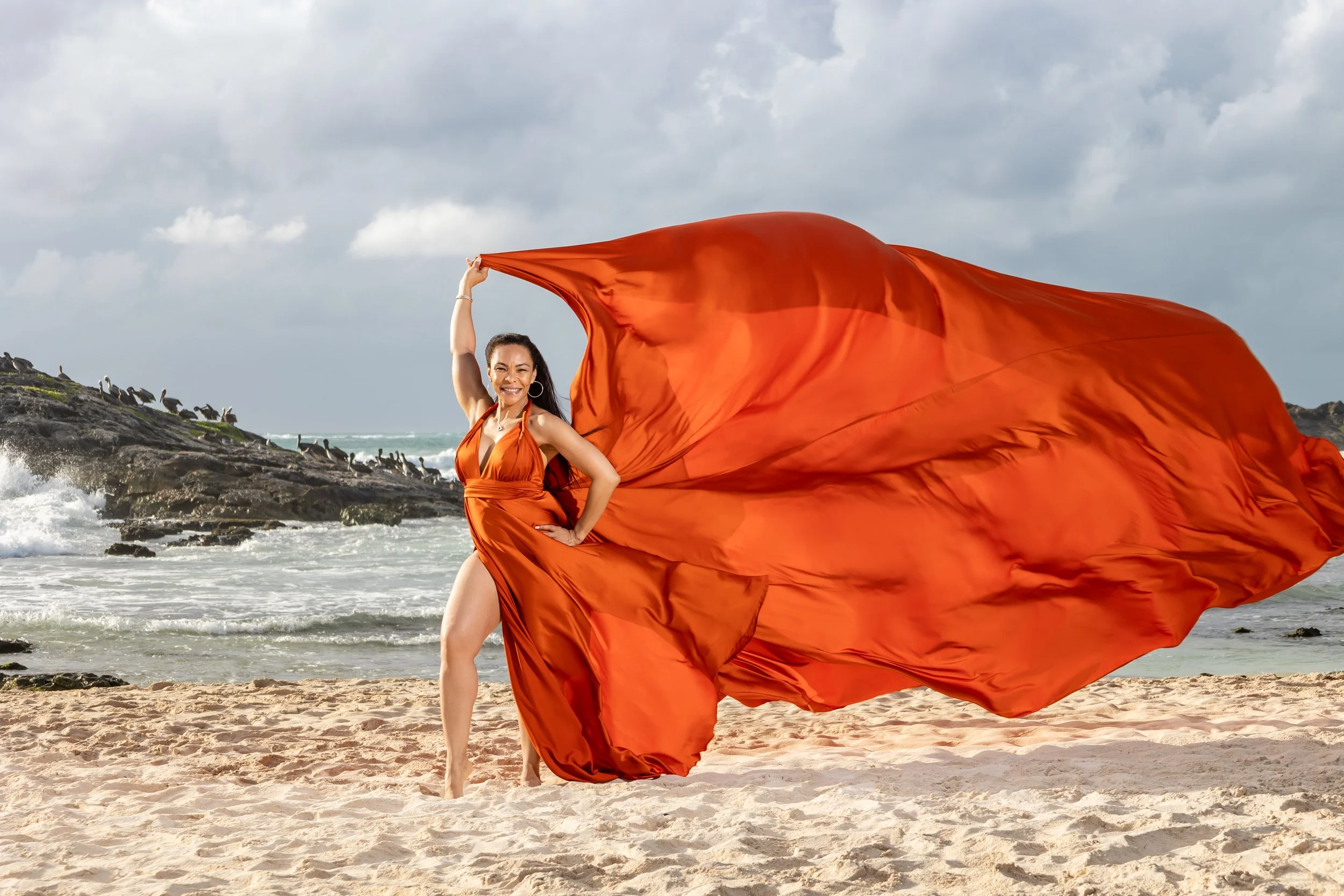 A woman in an orange gown and crown standing on a beach by the water, facing away, with her skirt flowing on the sand.