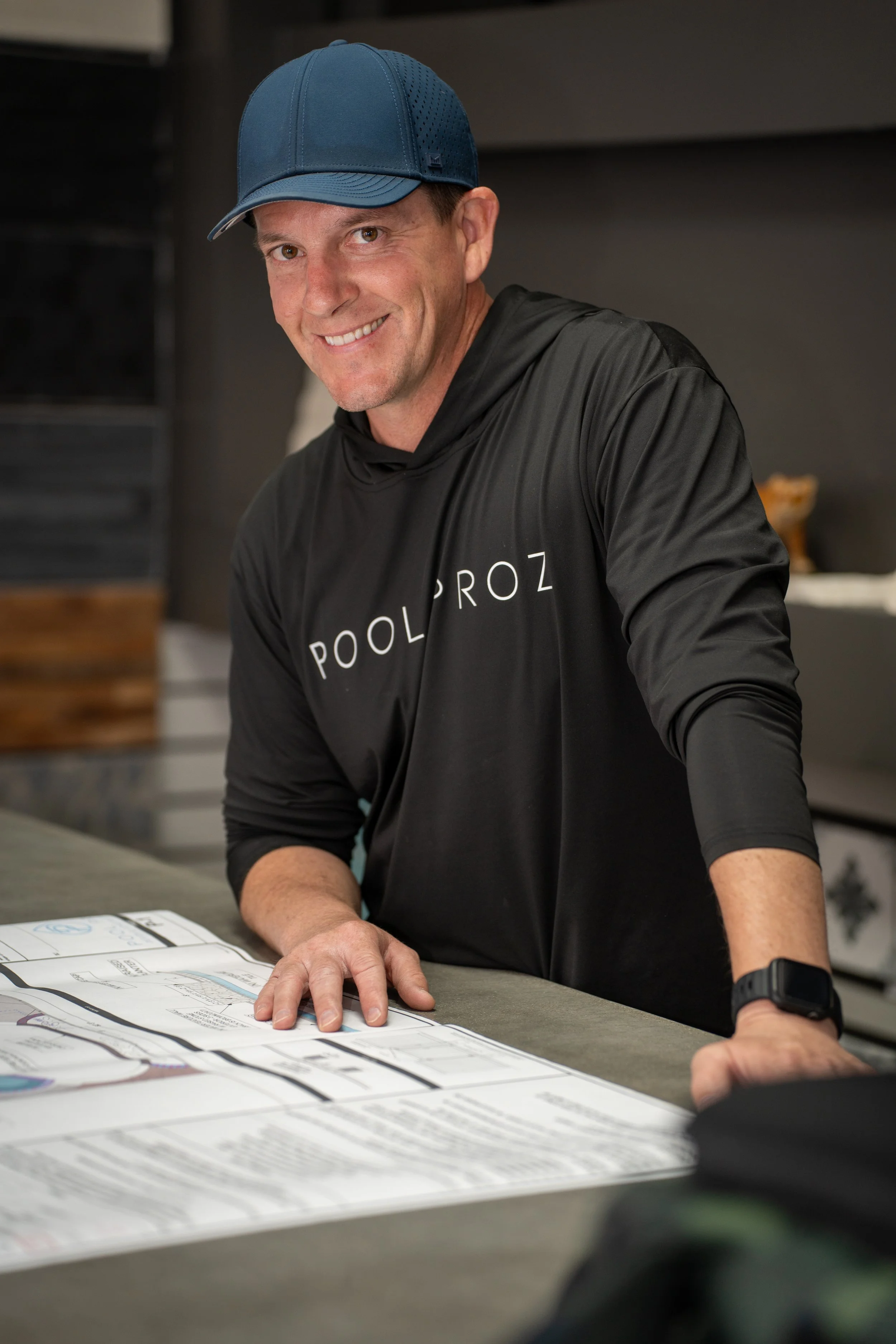 A smiling man wearing a blue cap and a black hoodie with 'POOLPROZ' printed on it, standing at a table with papers in what appears to be a pool or game room.