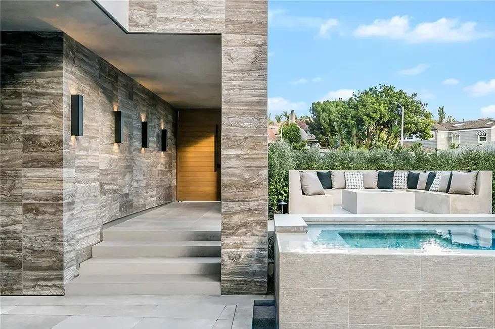 Modern outdoor patio with a pool, built-in seating, and a stone wall with black wall sconces, under a clear blue sky.