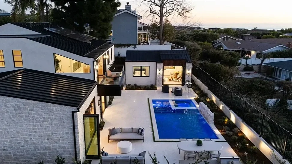 A modern backyard with a swimming pool and hot tub, outdoor seating, and a view of neighboring houses and trees at sunset.