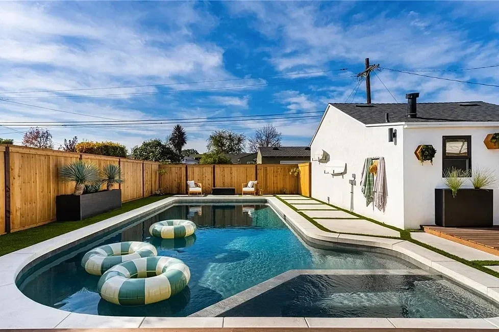 A backyard with a swimming pool, lounge chairs, potted plants, and a white house with hooks for hanging towels. The sky is partly cloudy with power lines overhead.
