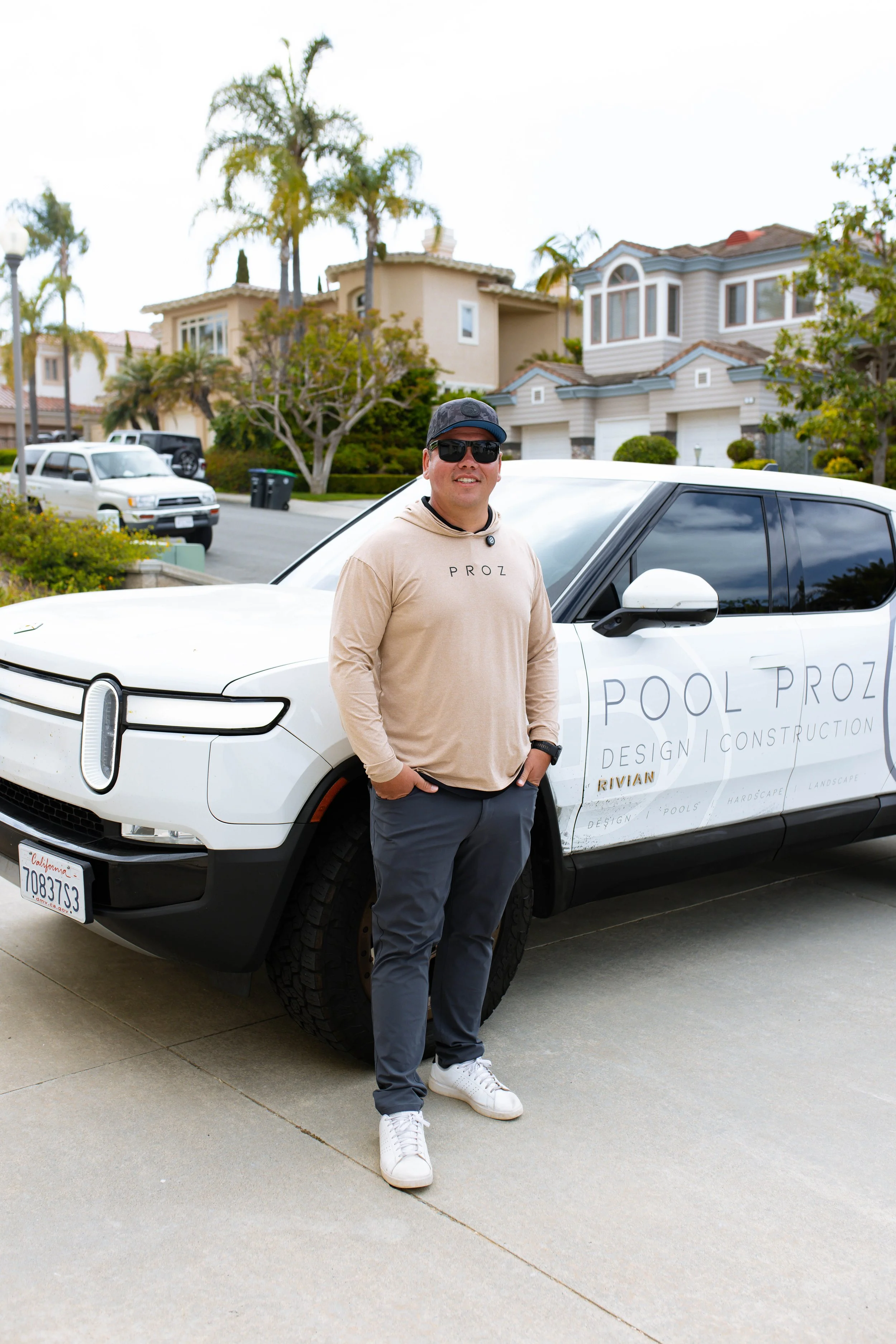 A man standing next to a white electric vehicle with 'Pool ProZ' logo parked on a suburban street. The man is wearing sunglasses, a black cap, a beige hoodie, and gray pants.