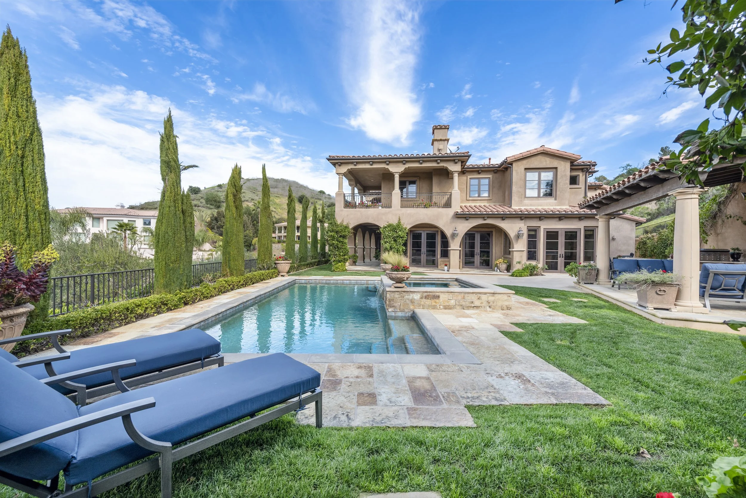 Backyard of a luxurious house with a swimming pool, lounge chairs, lush green grass, tall trees, and a patio area with seating, under a partly cloudy sky.