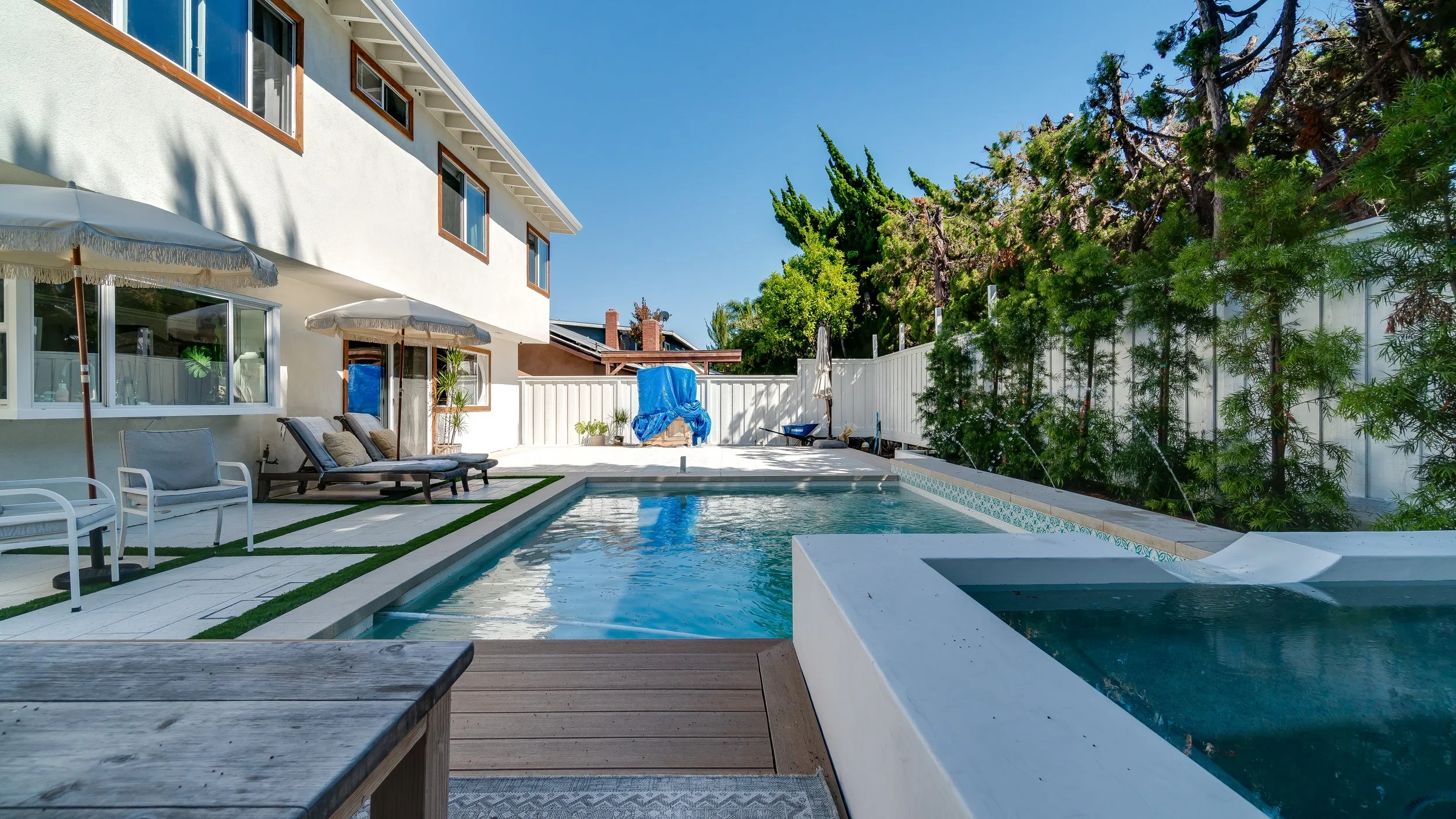 Backyard with a swimming pool, lounge chairs with cushions, umbrellas, and trees along the fence under a clear blue sky.