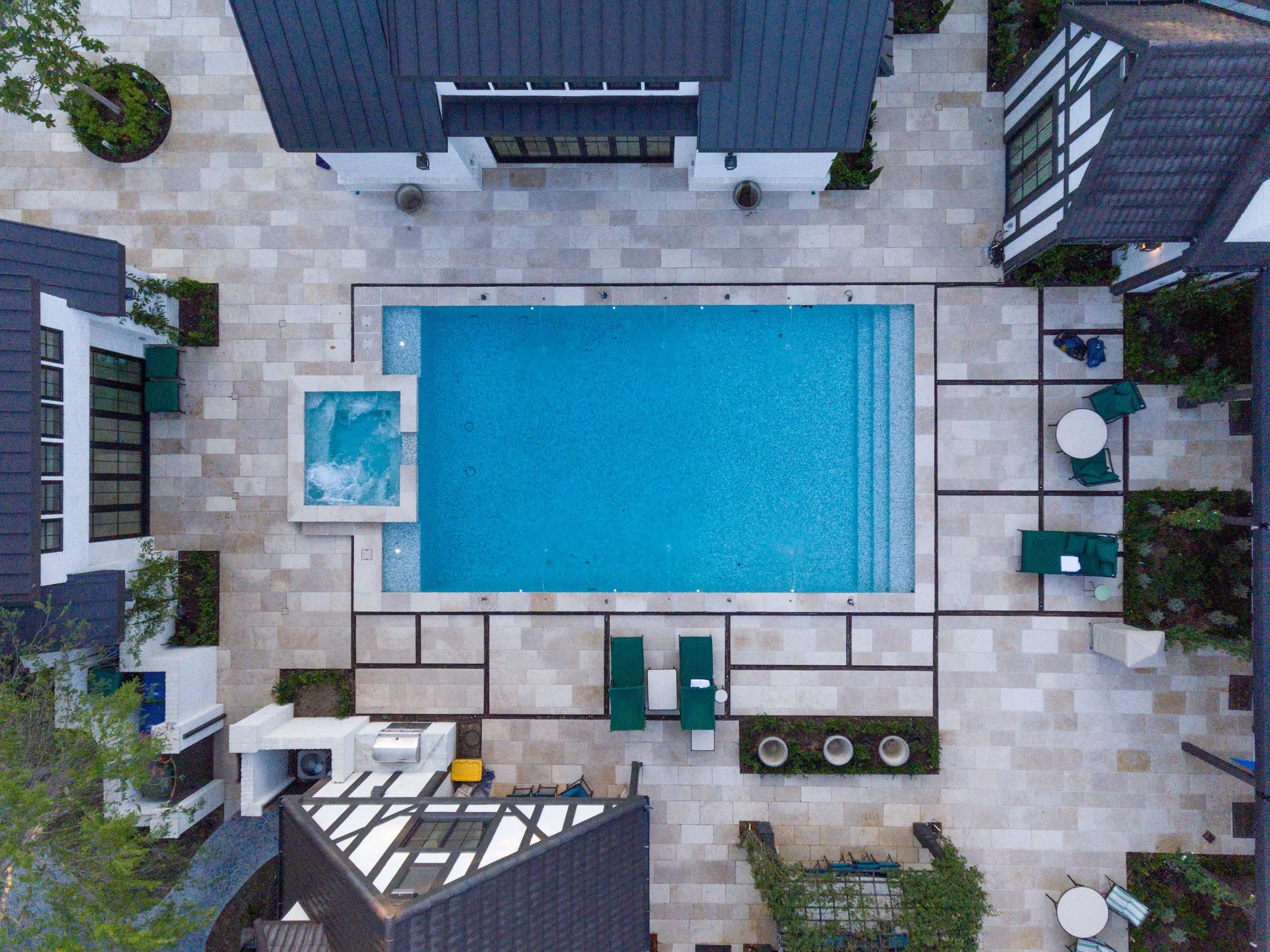 An aerial view of a modern swimming pool area in a residential backyard. The pool is rectangular with a small attached hot tub at one end. Surrounding the pool are tiled patios with outdoor furniture, including chairs, tables, and lounge chairs. Ther