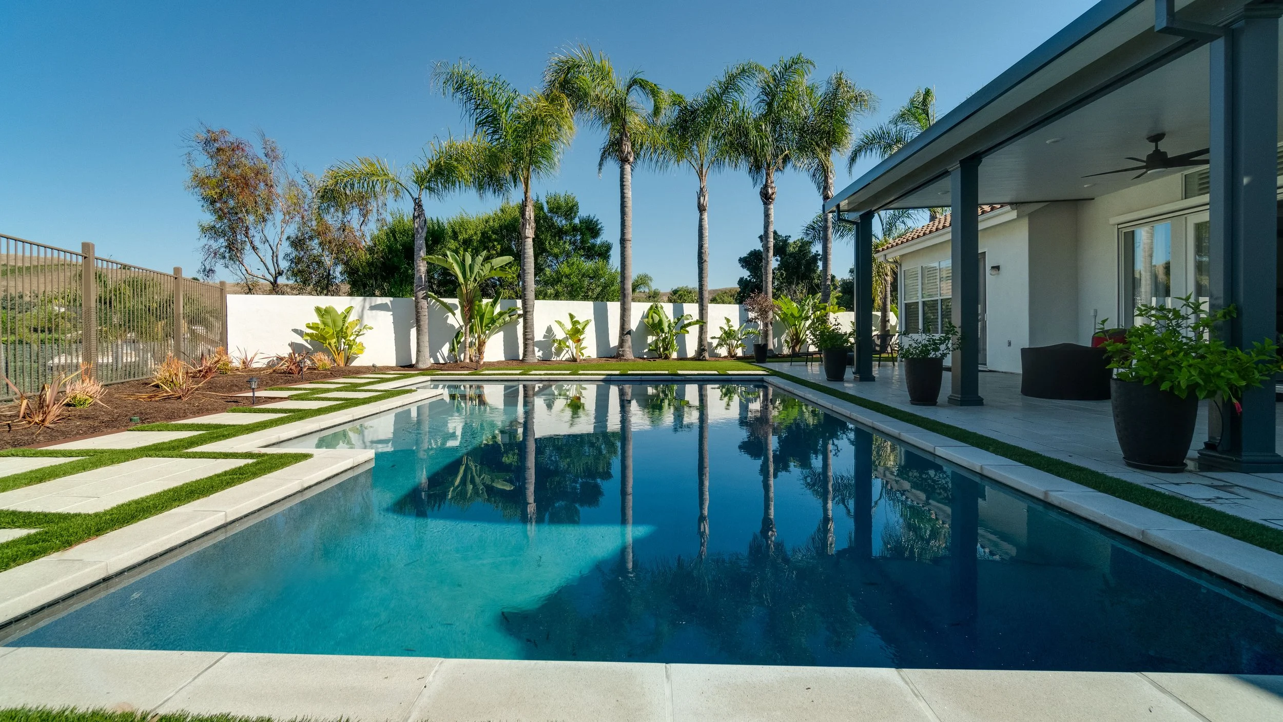 Backyard with a rectangular swimming pool, palm trees, potted plants, and a shaded patio area with a ceiling fan and outdoor furniture.