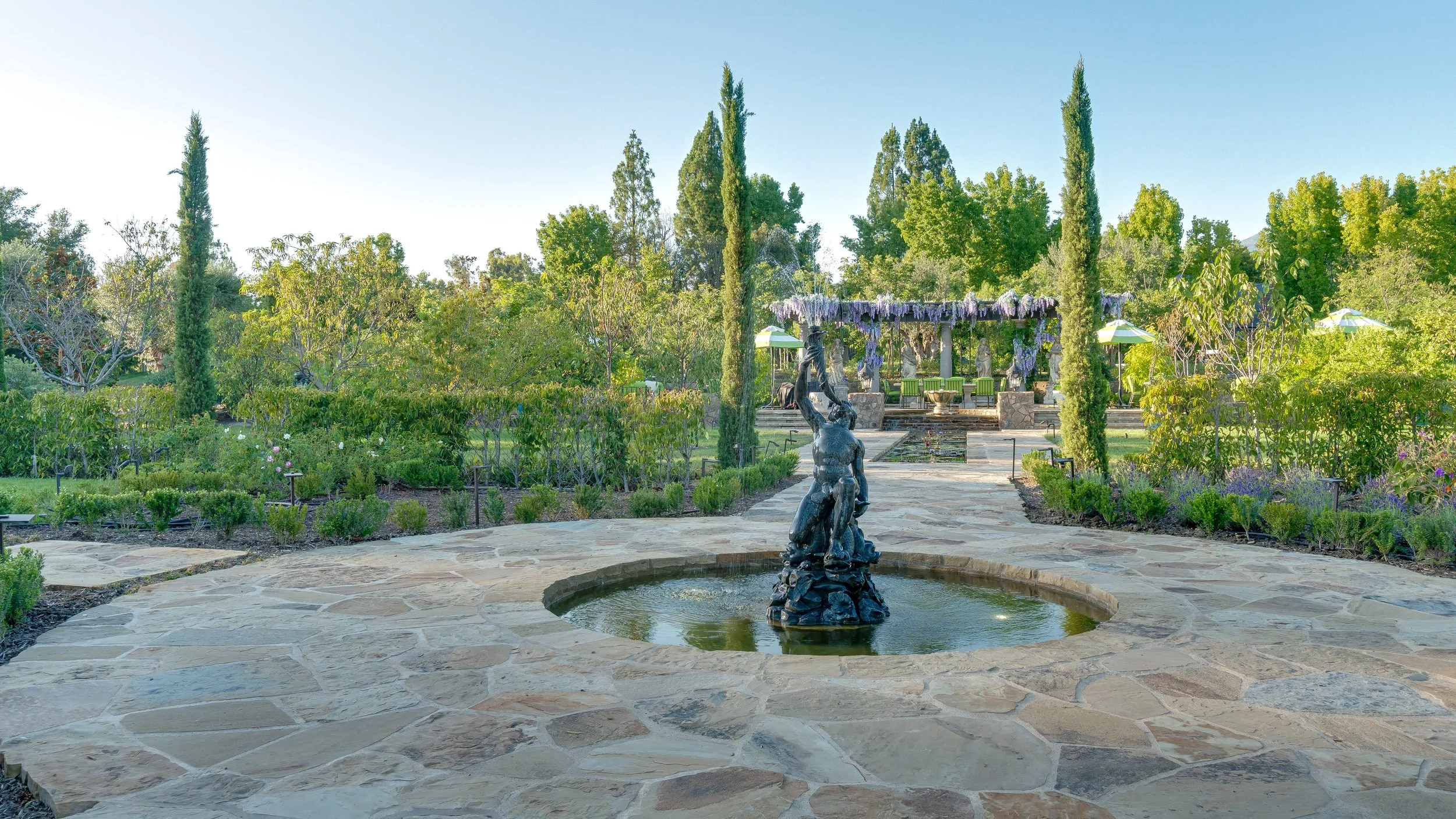A landscaped garden with a stone pathway, a fountain centerpiece with a statue of a woman holding a vessel, lush greenery, tall cypress trees, and a pergola with purple hanging flowers in the background.
