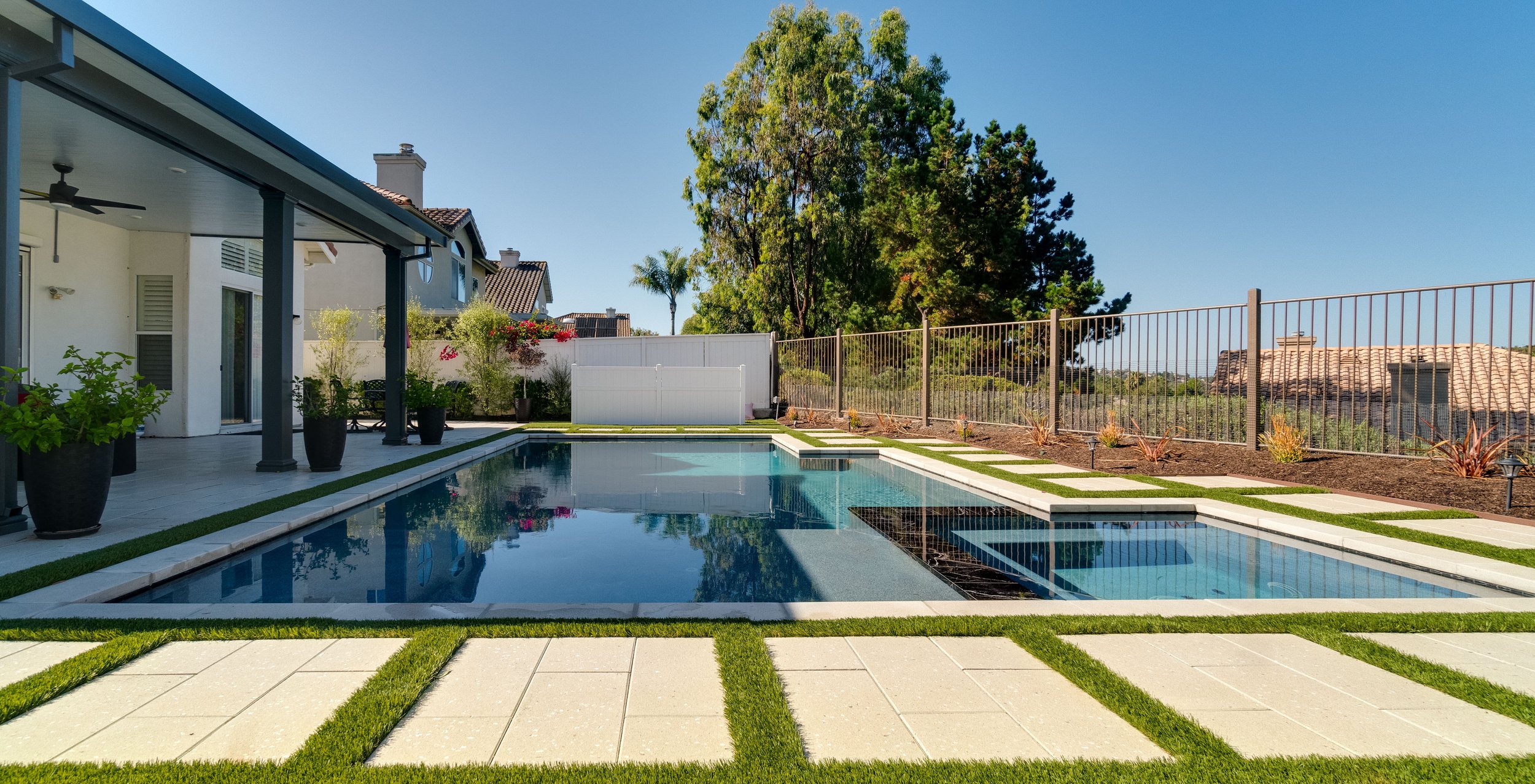 Backyard with a swimming pool, potted plants along the patio, a fence on the right side, trees in the background, and neighboring houses with tiled roofs under a clear blue sky.