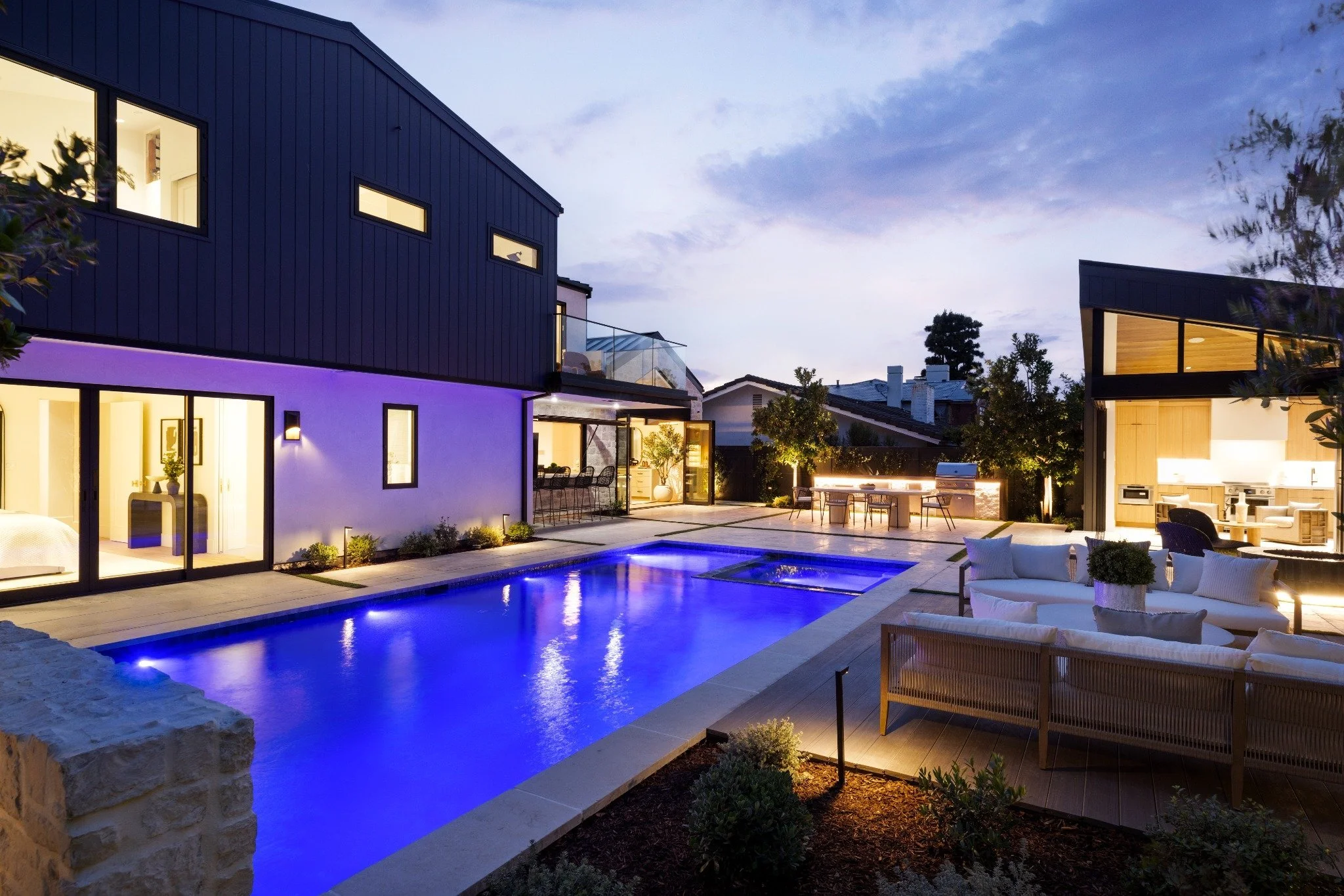 Modern house with illuminated interior and outdoor pool area at dusk, featuring outdoor seating, dining area, and kitchen.