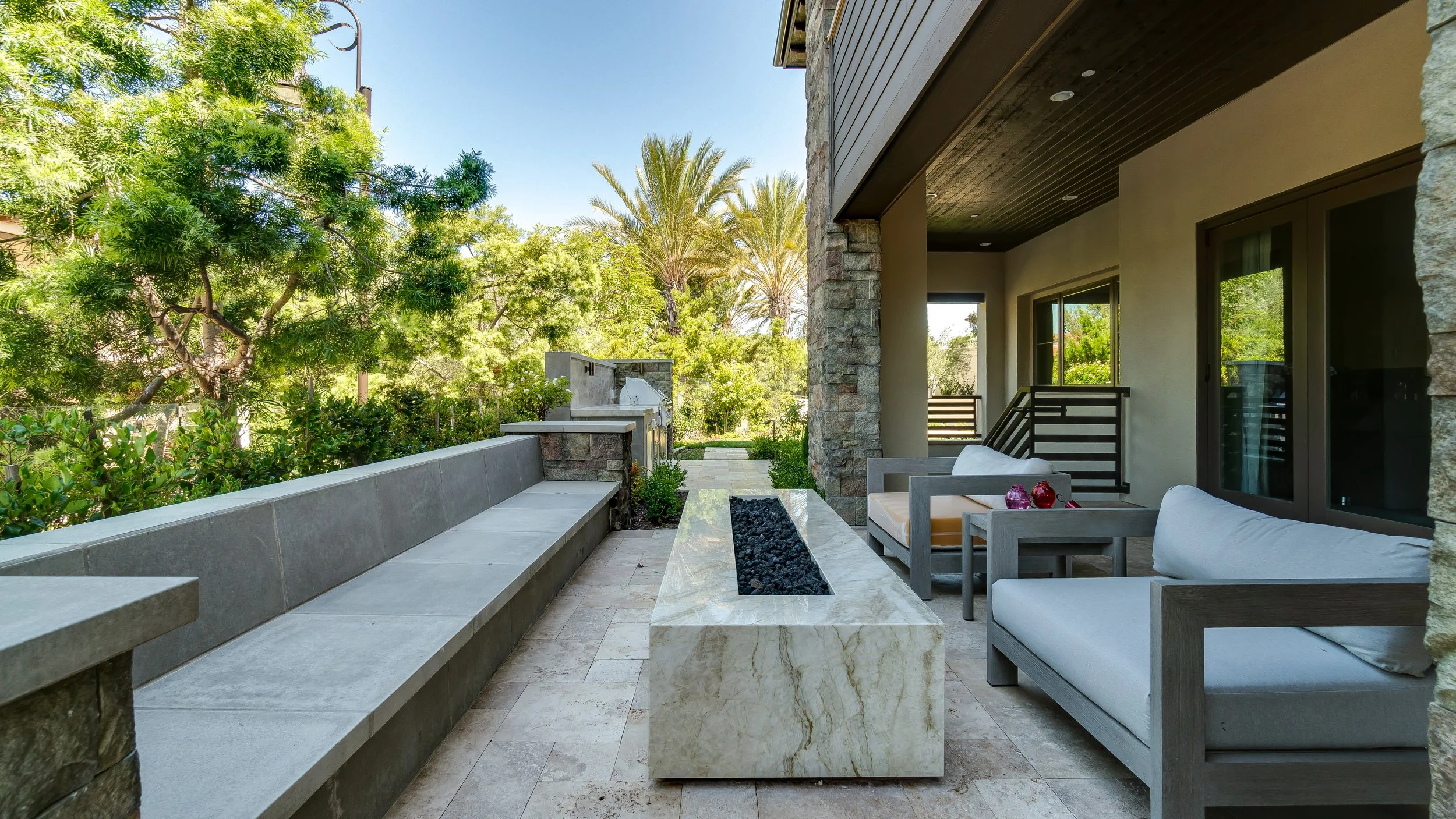 Outdoor patio with modern furniture, greenery, trees, and a built-in fireplace or grill area.