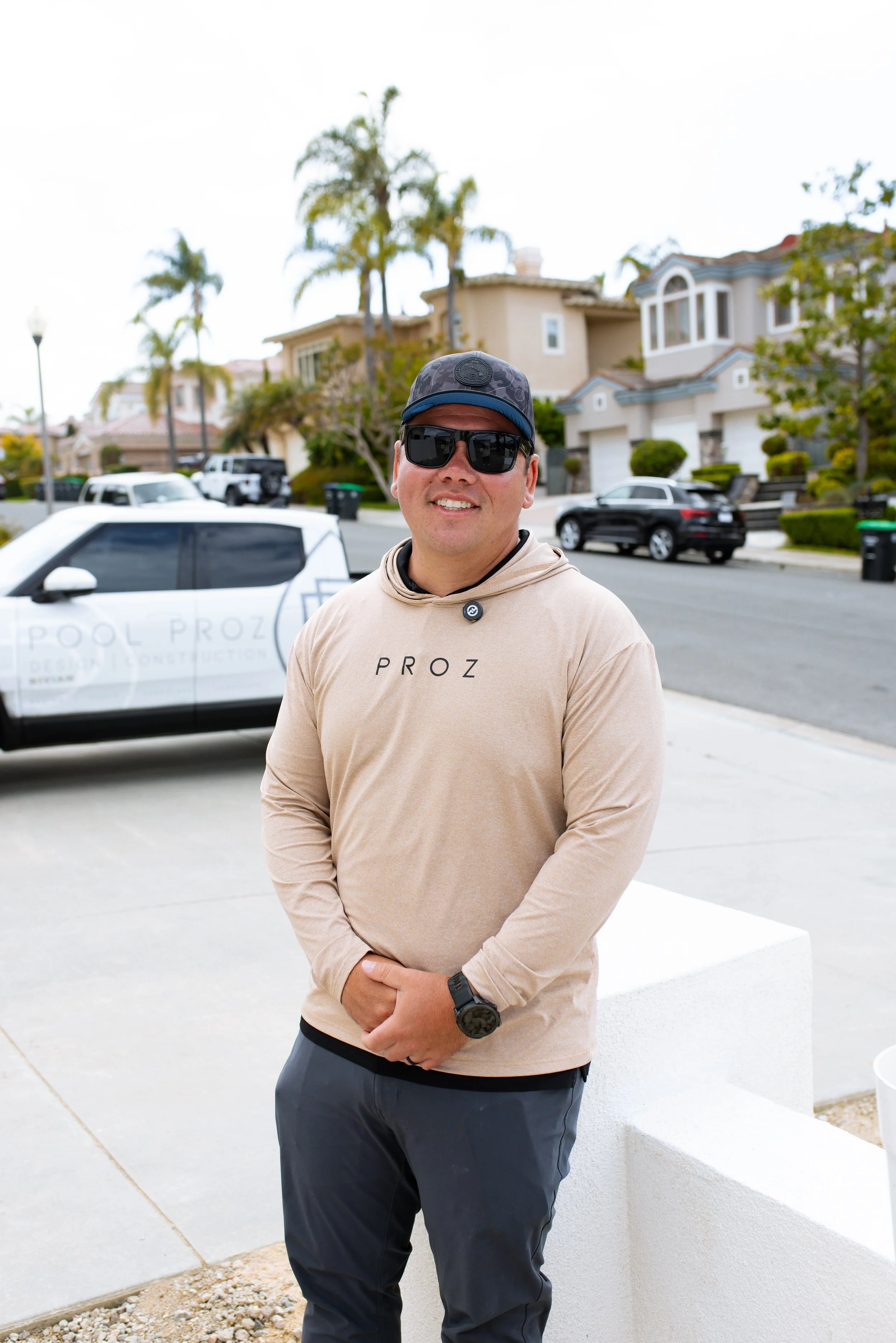 A smiling man wearing sunglasses, a beige hoodie with 'PROZ' written on it, and a black cap, standing outdoors in front of a residential neighborhood with palm trees and houses.