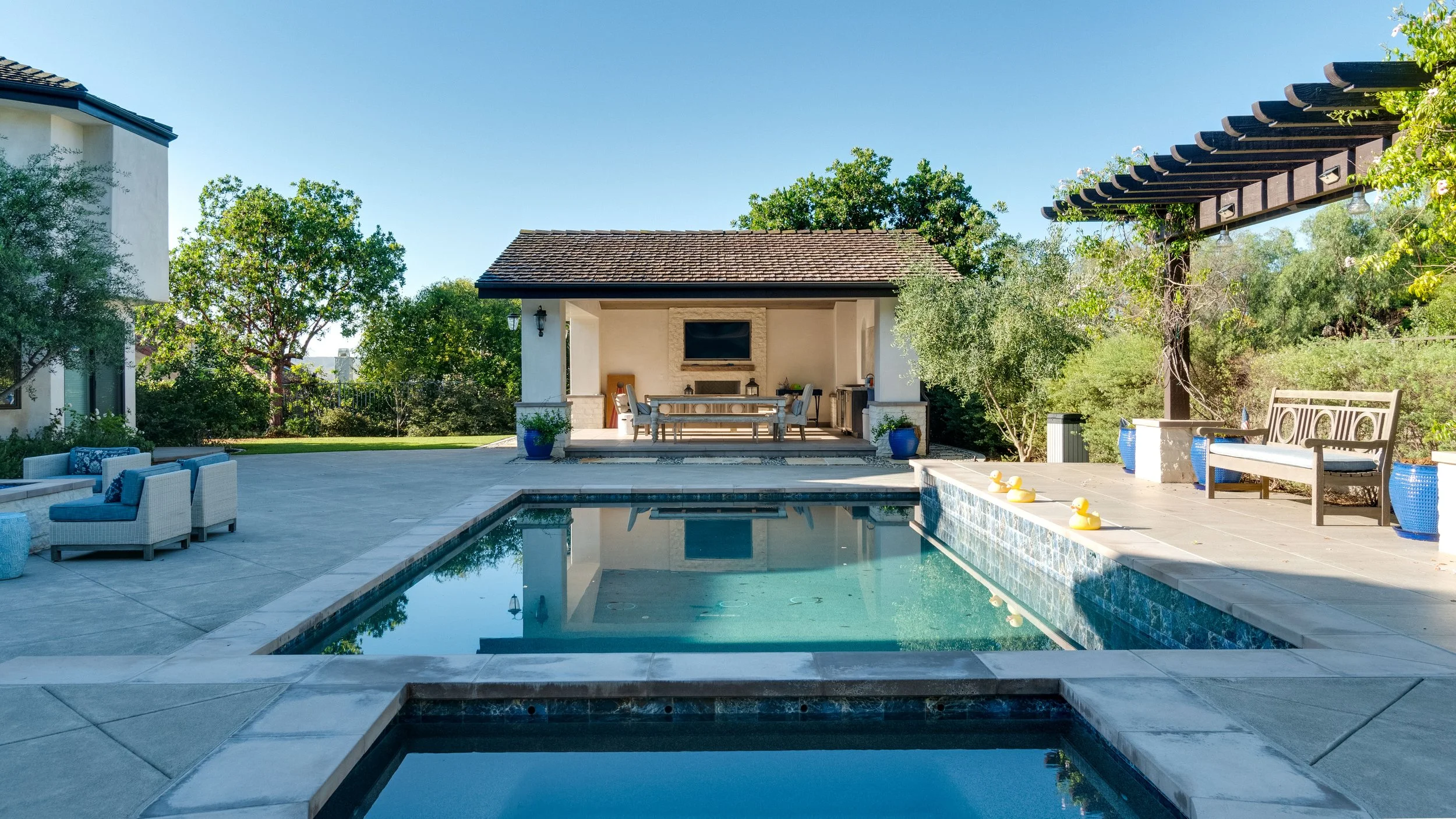 A backyard with a swimming pool, outdoor seating, and a small pavilion with a television, surrounded by trees and greenery.