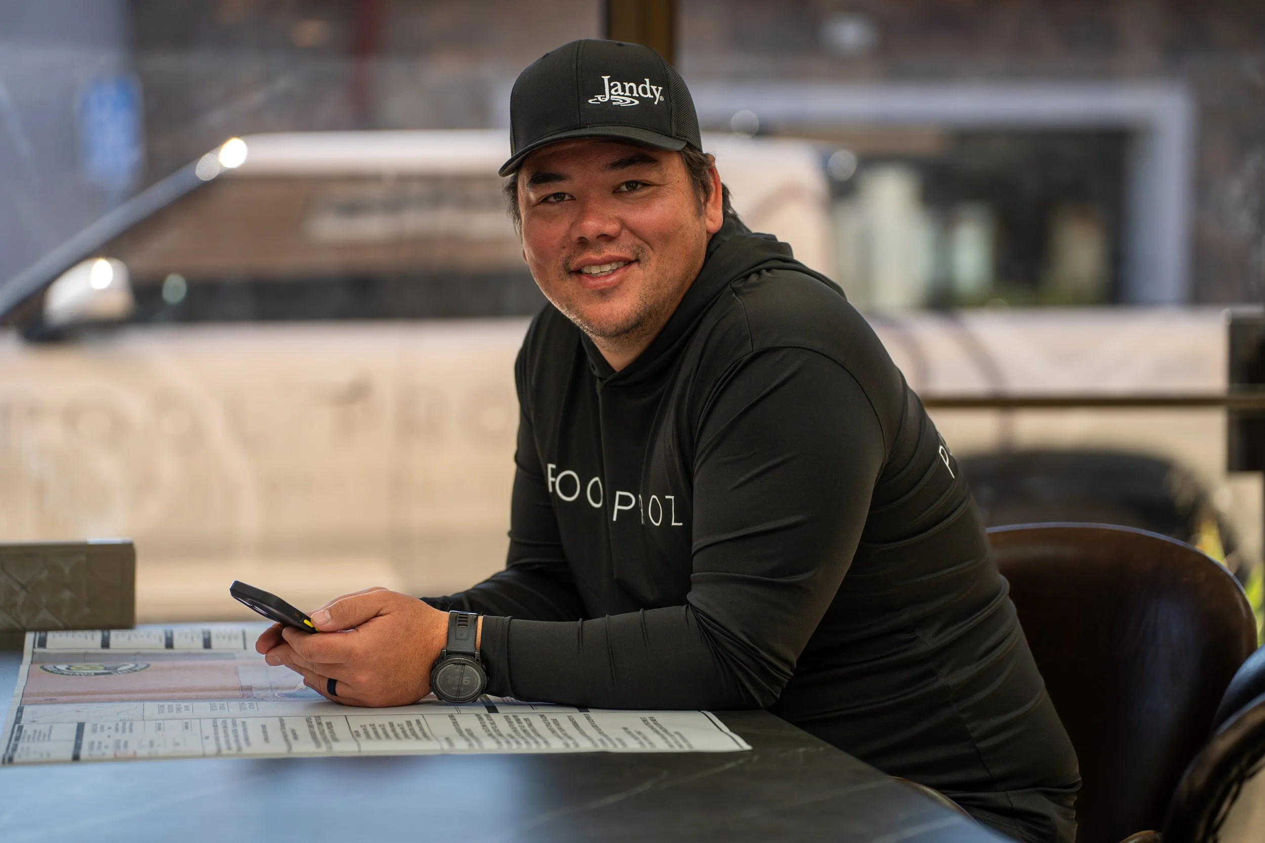 A man sitting at a table in a restaurant or cafe, wearing a black JandY cap and black hoodie, smiling and holding a phone, with a menu on the table.