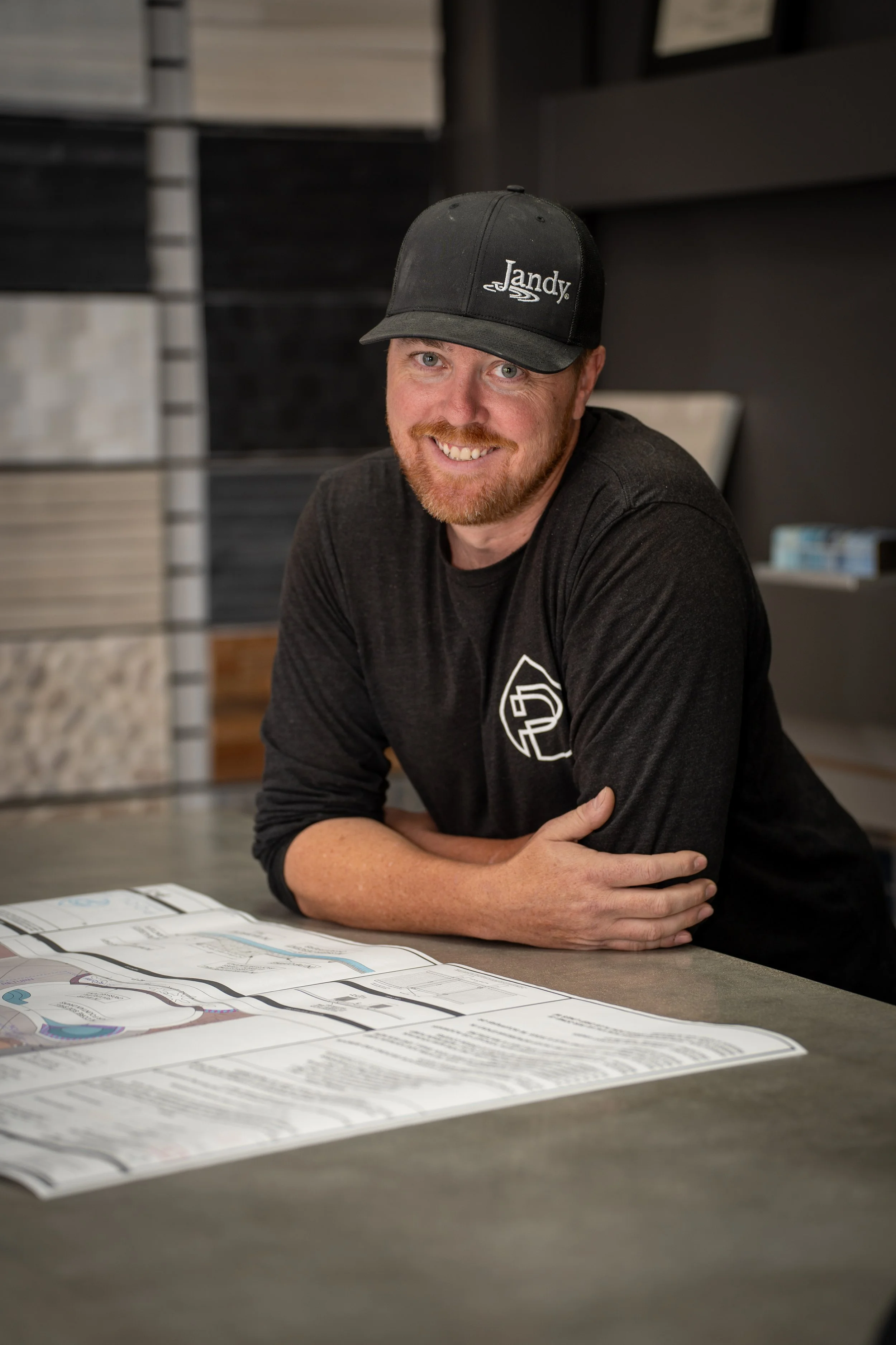 A man smiling and wearing a black cap and black long-sleeve shirt, sitting at a table with architectural blueprints or plans.