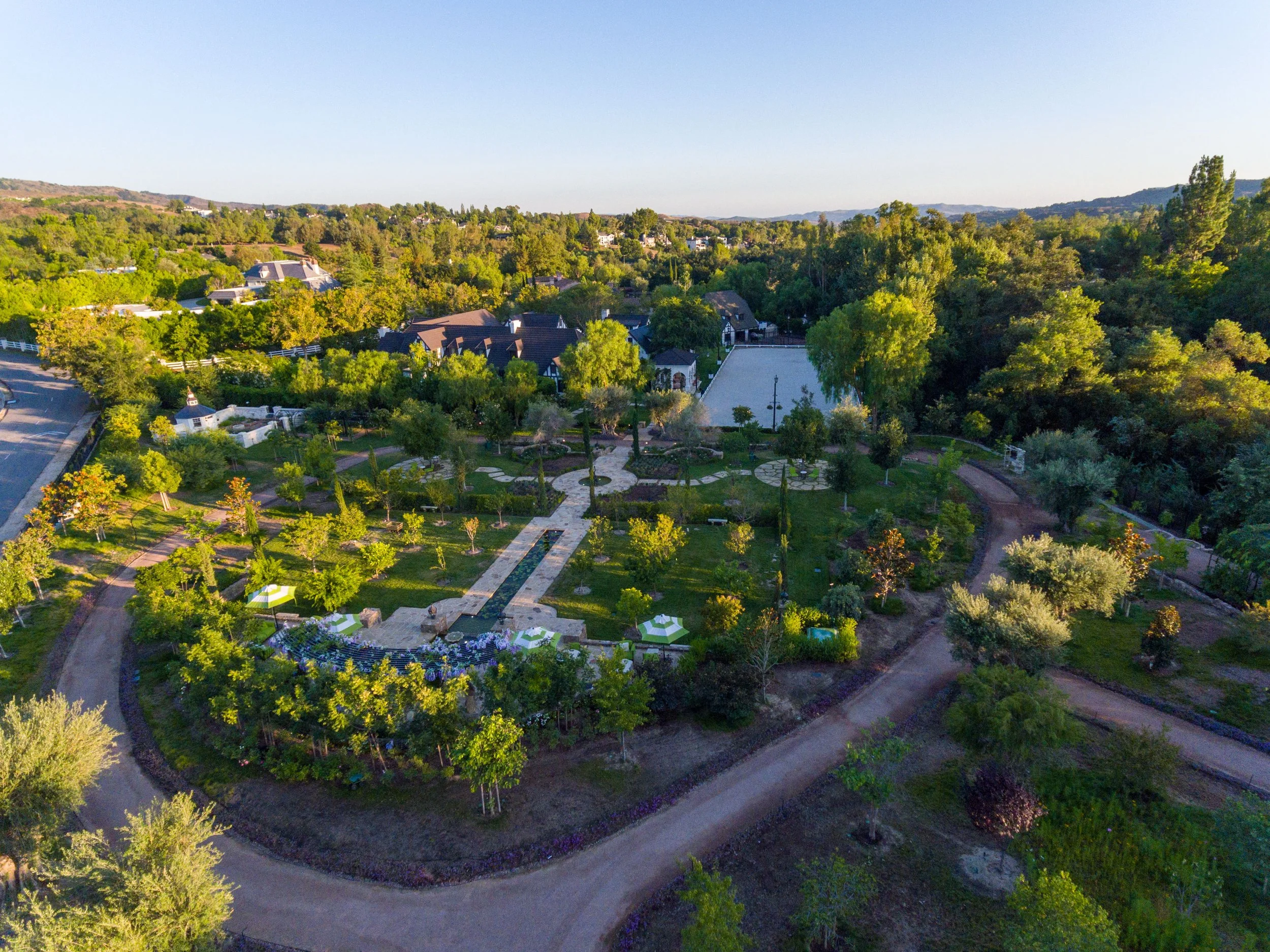 An aerial view of a lush park with walking paths, green trees, a small garden area, and a tennis court, surrounded by houses and roads in the background, under a clear sky.