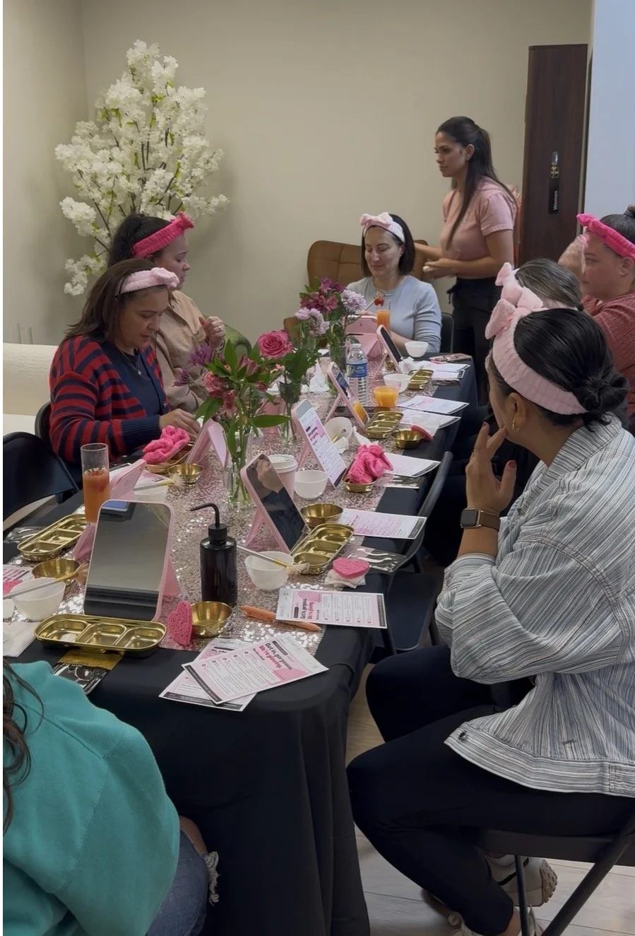 Women sitting at a decorated table with pink and gold accents, pink headbands, and pink plush slippers, participating in a gathering or spa event, with a white flower arrangement in the background.