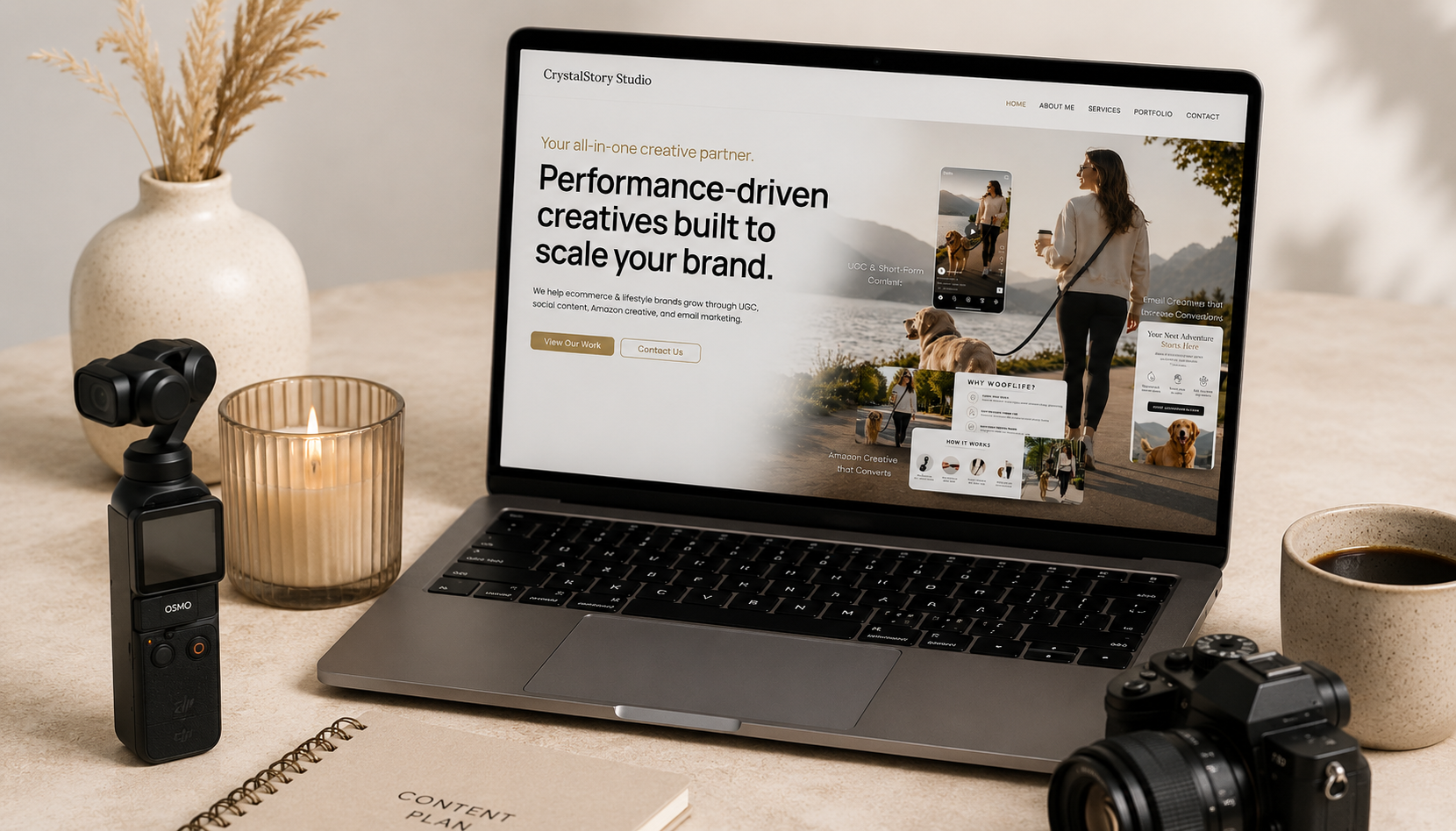 Laptop displaying a website with the headline 'Performance-driven creatives built to scale your brand.'; to the left, a camera mounted on a stabilizer, a lit candle, and a vase with dried pampas grass; on the right, a cup of coffee, a camera, and a content planner notebook.