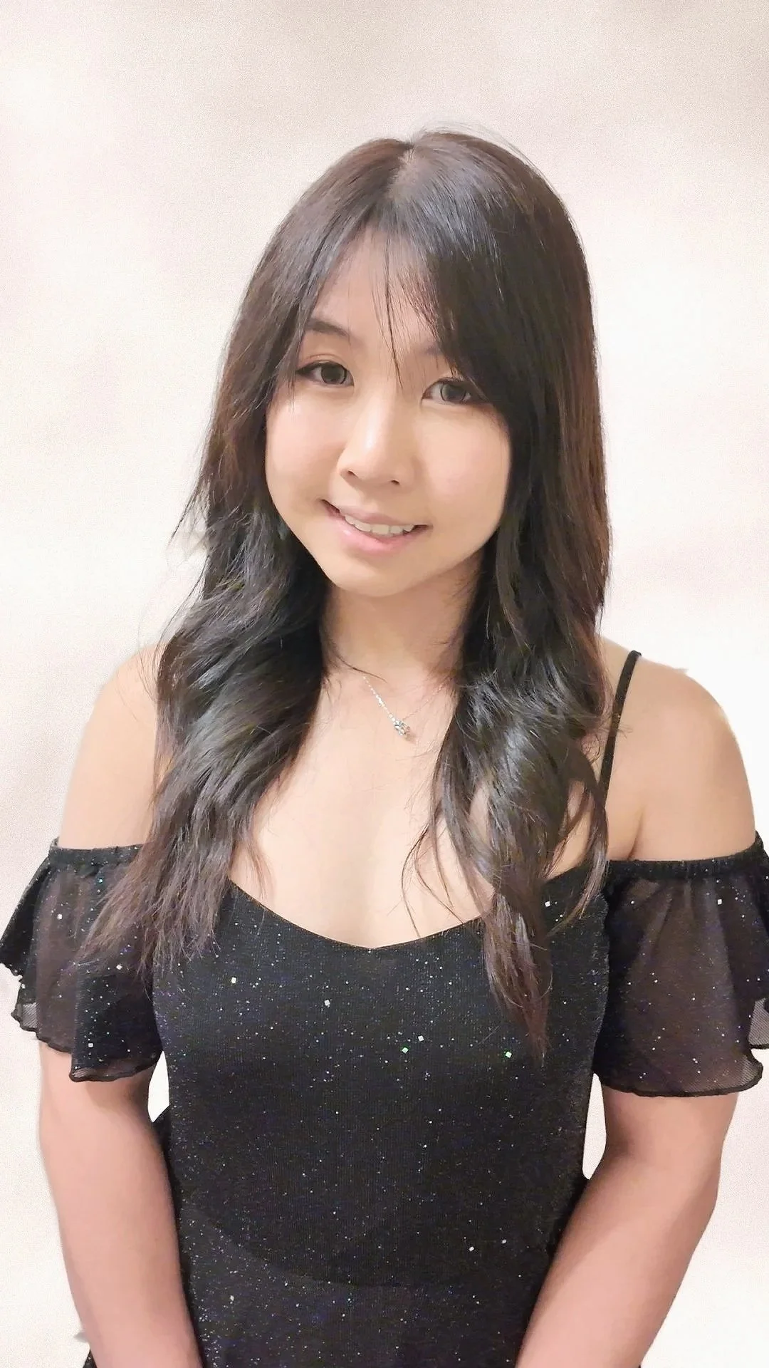 A young woman with dark wavy hair, wearing a black sparkly off-shoulder top with spaghetti straps, smiling at the camera against a plain light background.