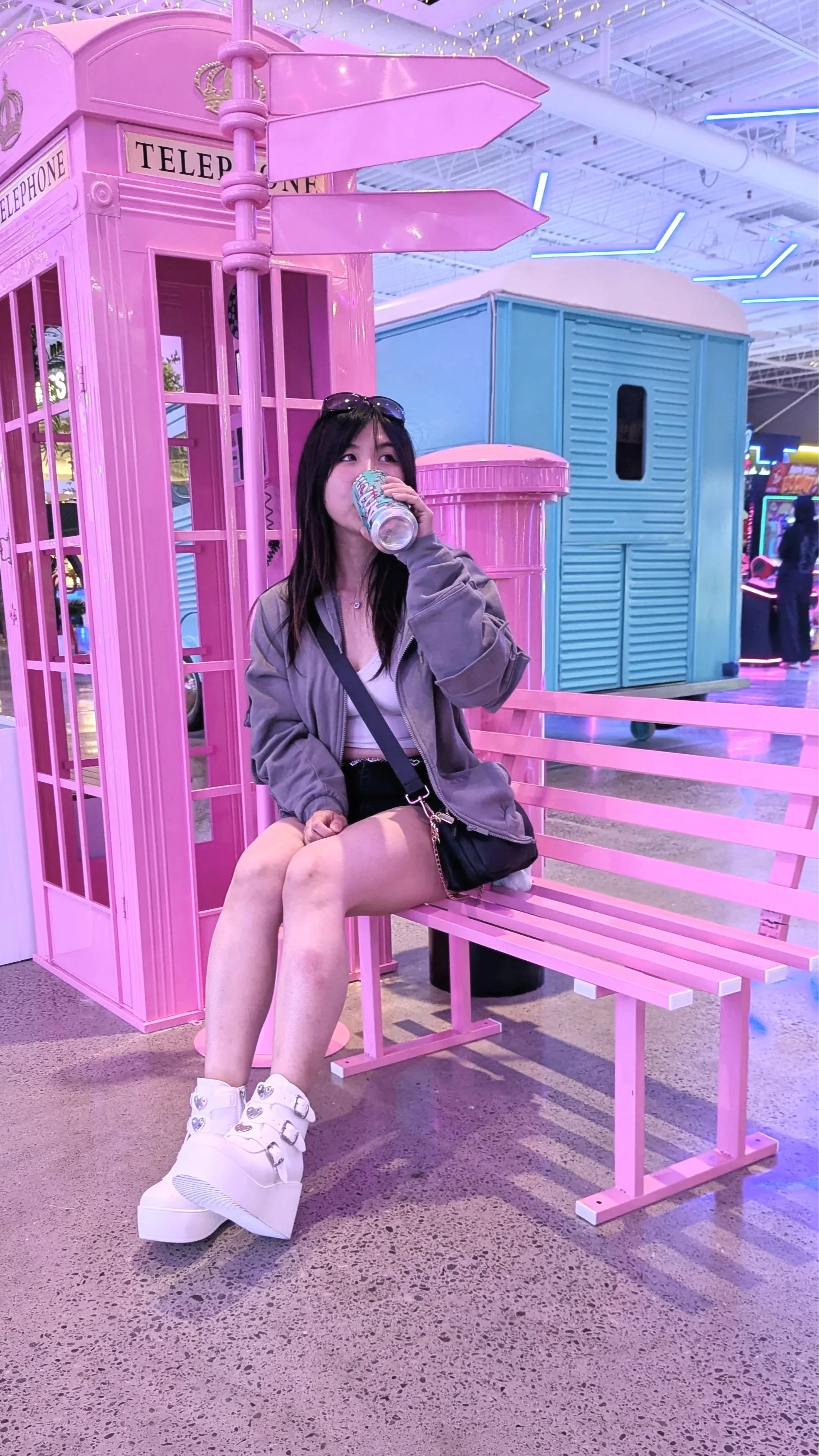 A young woman with long dark hair, wearing a grey jacket, white top, black shorts, and white platform shoes, sits on a pink bench in a colorful indoor arcade or amusement park. She is drinking from a can and has sunglasses on her head, with a small black bag hanging from her shoulder. Behind her are pink and blue decorative structures, including a pink phone booth and a pink signpost.