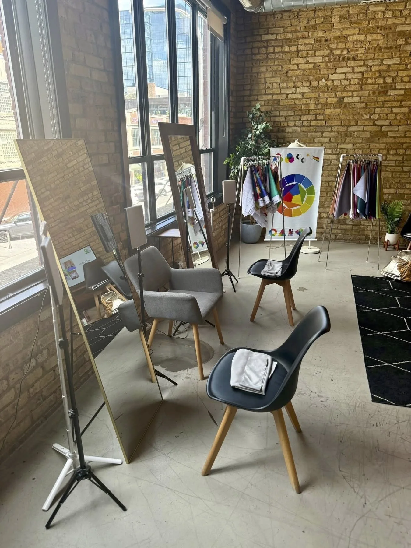 A modern interior space with large windows, brick walls, and a variety of chairs, mirrors, and clothing displays, suggesting a creative workspace or boutique.