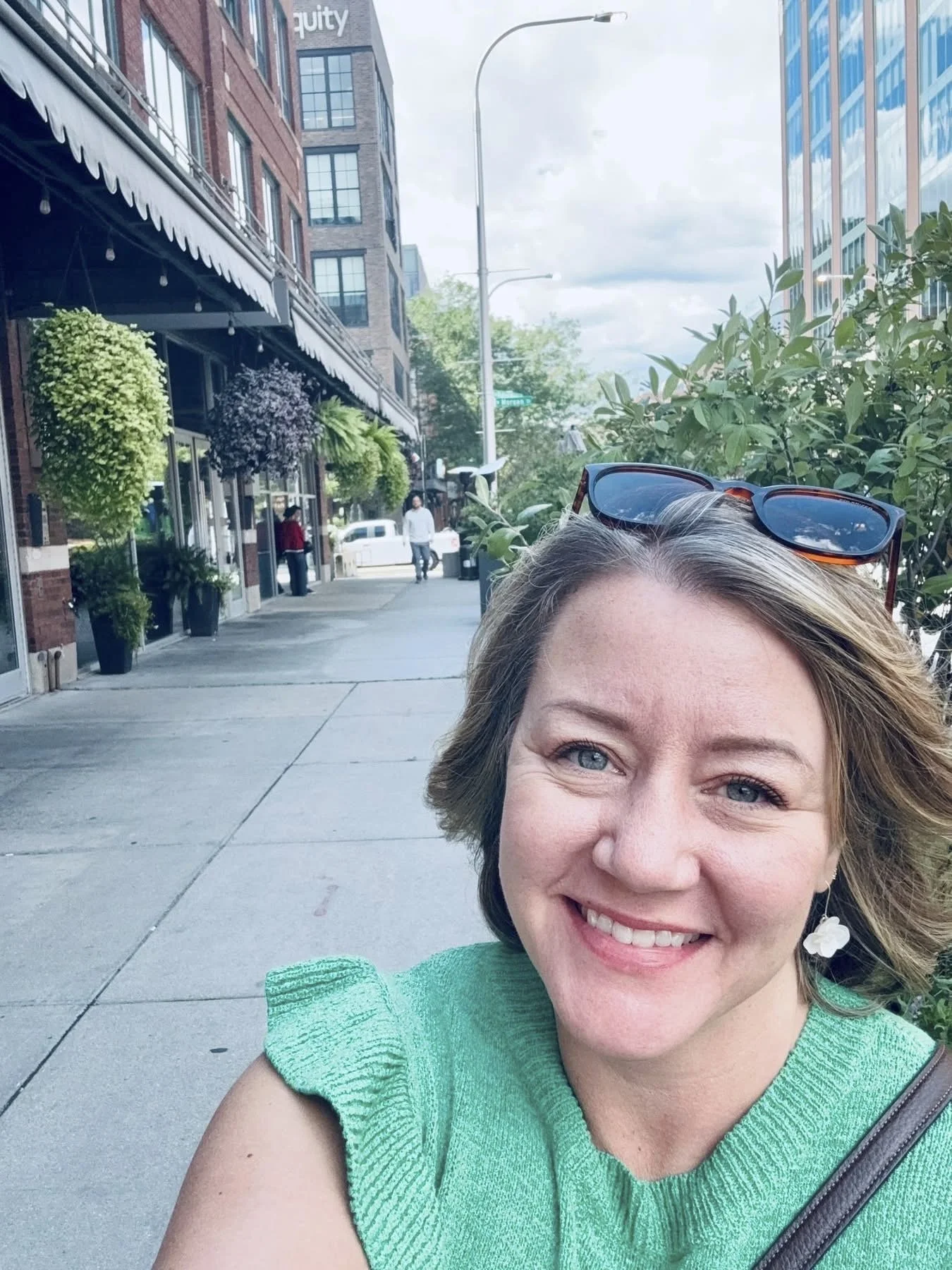 A woman with short gray hair and blue eyes taking a selfie outside on a city sidewalk, wearing a green top and sunglasses on her head, with buildings and plants in the background.