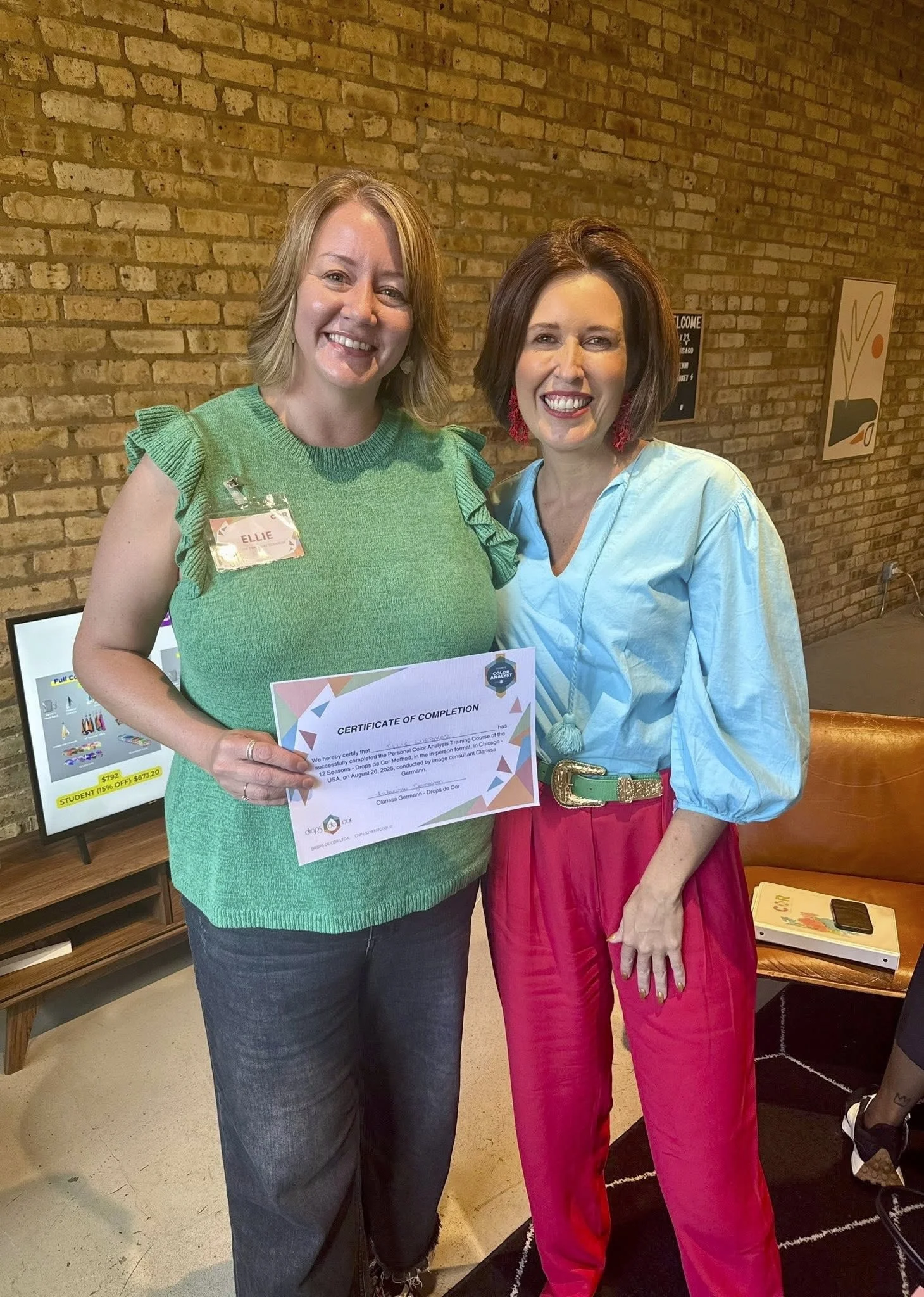 Two women standing side by side smiling at the camera. The woman on the left is holding a Certificate of Completion, wearing a green top with ruffled sleeves and dark jeans. The woman on the right has dark hair, wearing a light blue blouse and bright