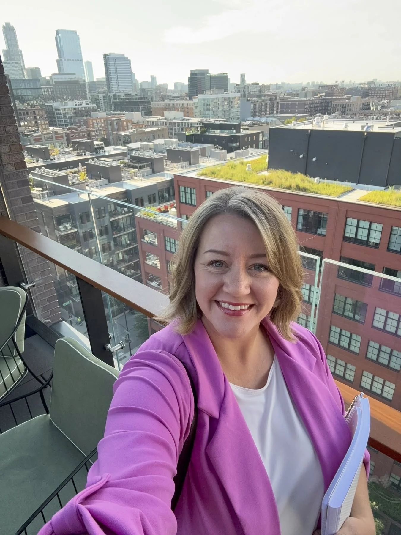A woman with shoulder-length blonde hair wearing a pink blazer and white top, smiling and taking a selfie on a balcony with a cityscape in the background.