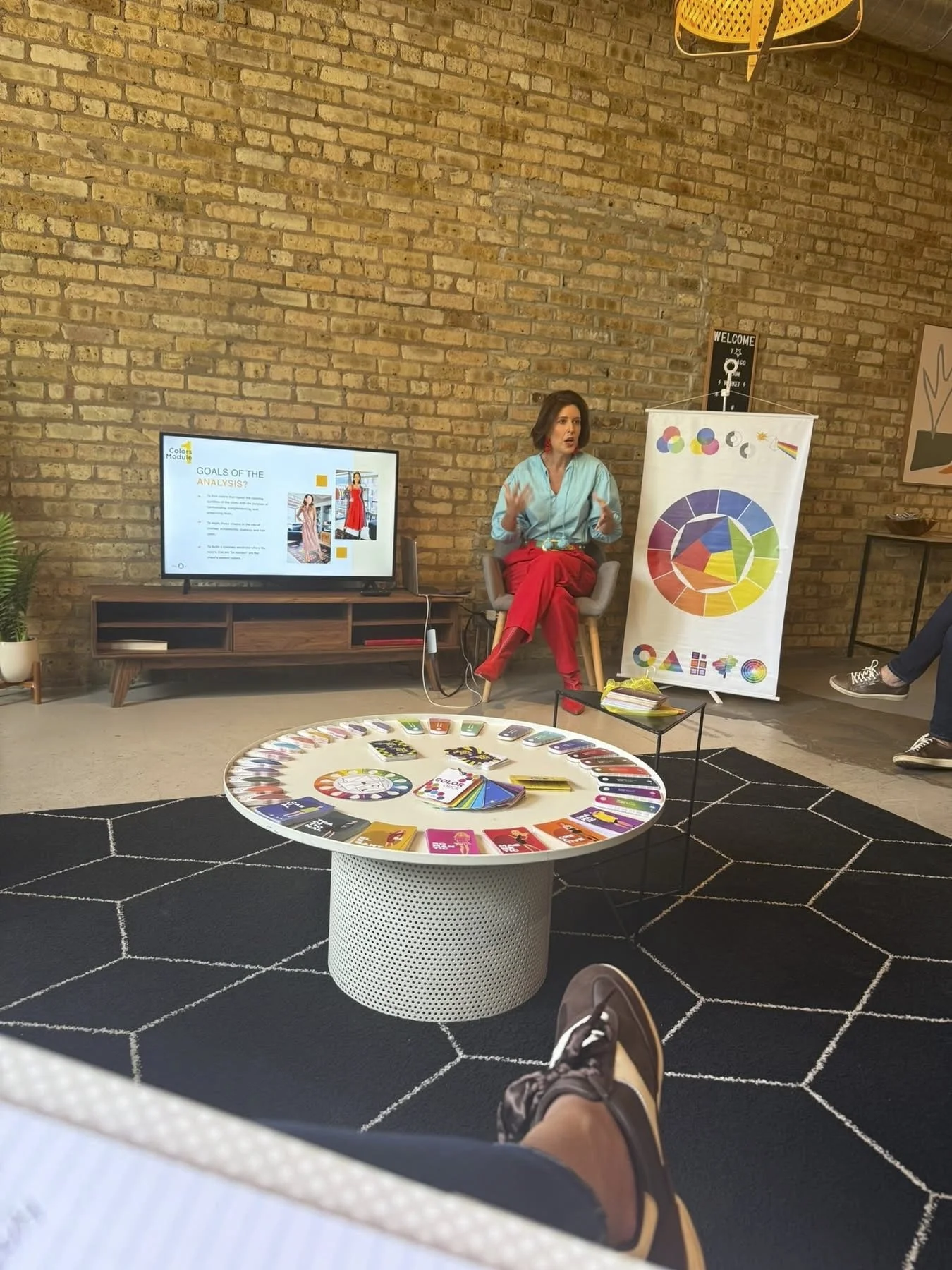 A woman giving a presentation in a modern, cozy room with a brick wall background. She is sitting on a chair, gesturing with her hands, and has a screen displaying presentation slides beside her. There is a colorful circular chart on a stand, a round