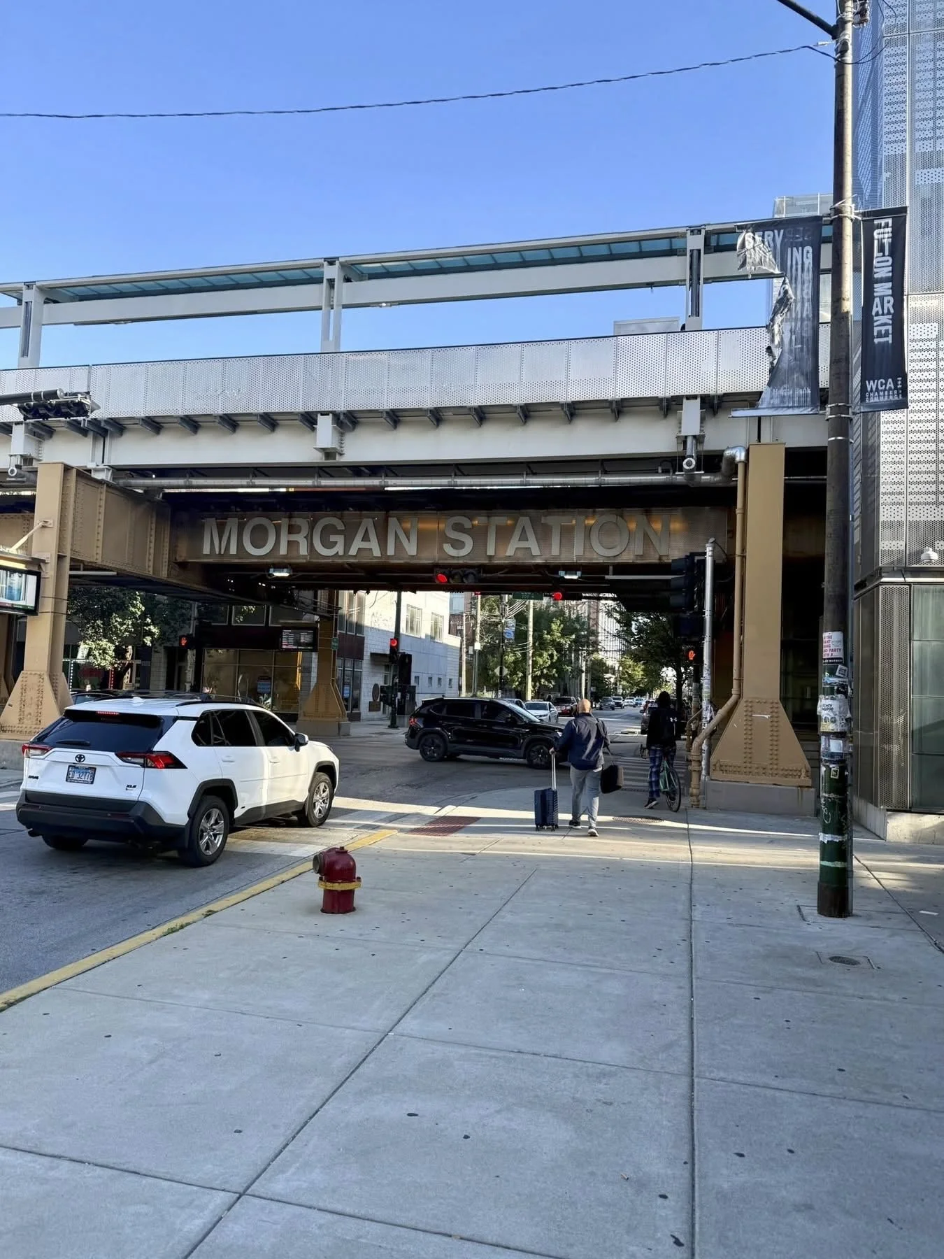 People walking on sidewalk with cars passing under elevated train track labeled Morgan Station in an urban area.