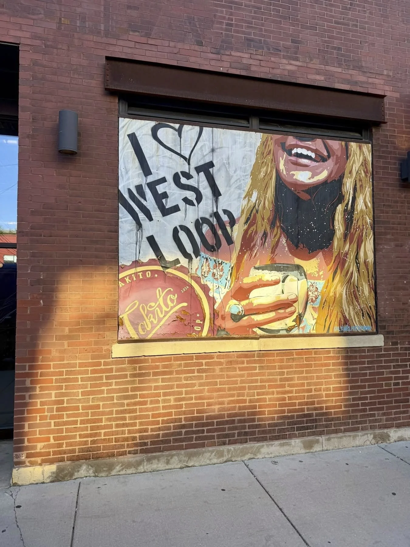 Street art mural on a brick wall showing a woman with long blonde hair, smiling and holding a drink, with a sign that says 'I love West Loop'.