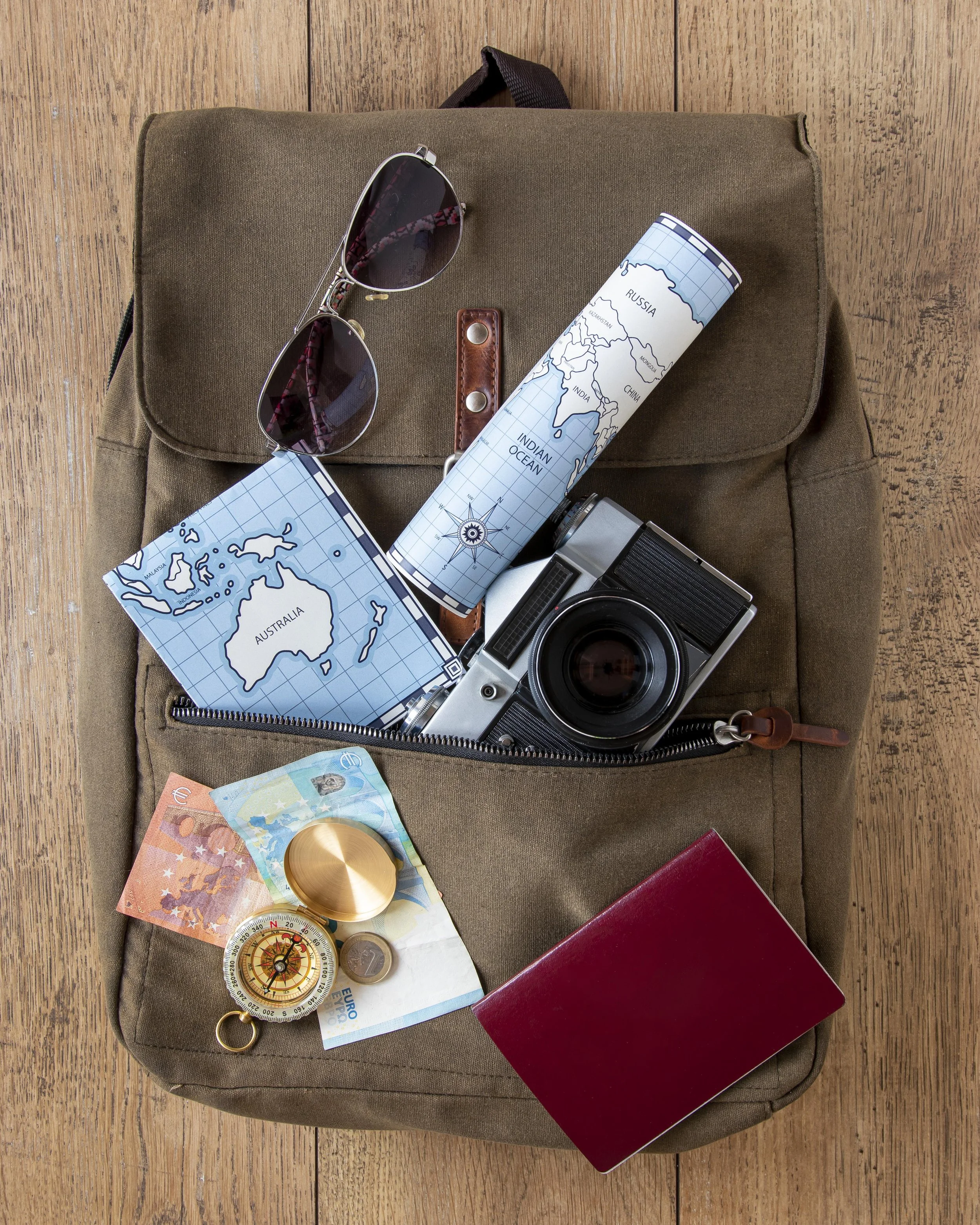 Travel backpack with sunglasses, maps, camera, passport, compass, and euros spilling out, flat lay on a wooden surface.