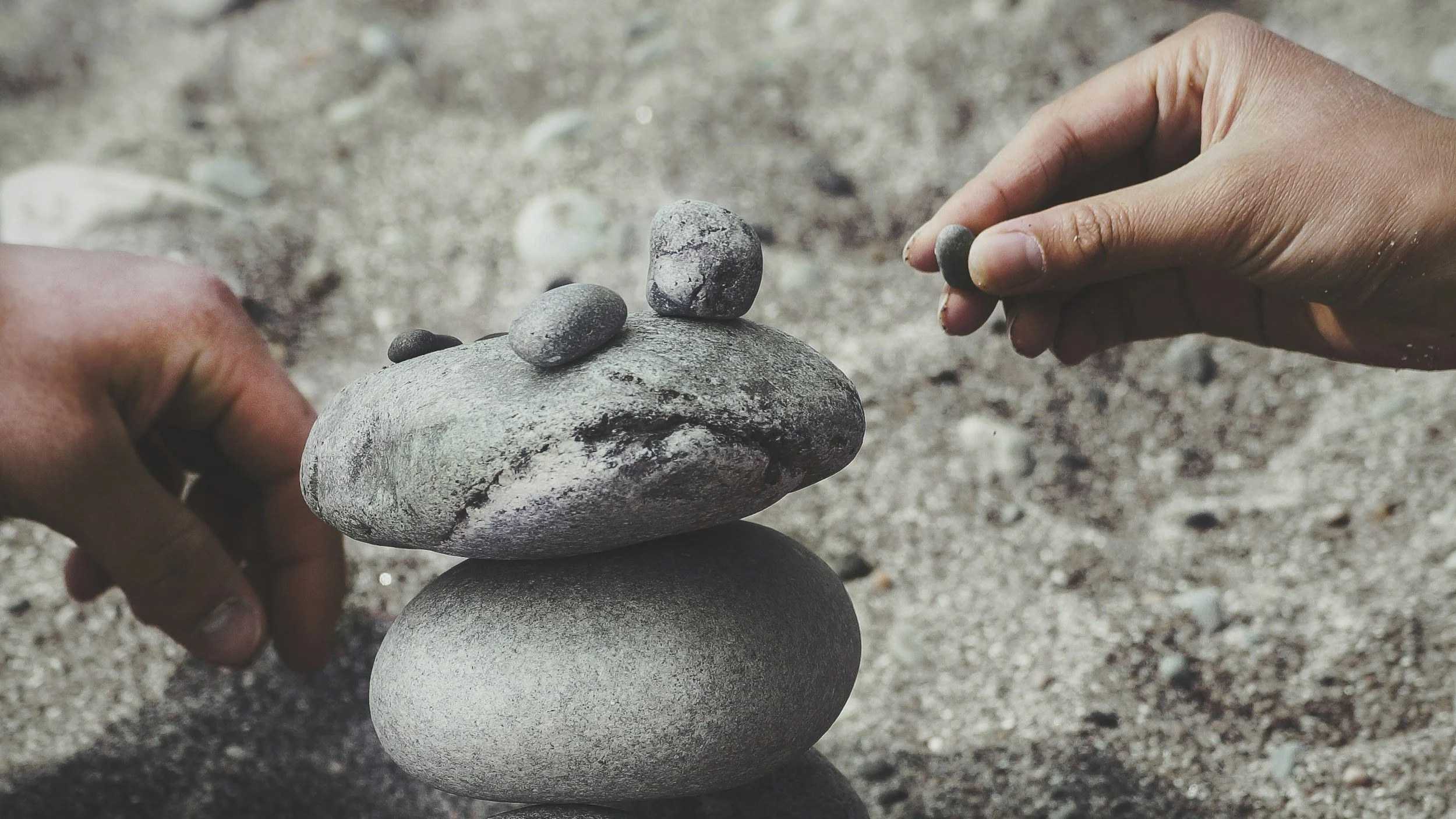 Hands stacking smooth, rounded stones on a sandy surface.